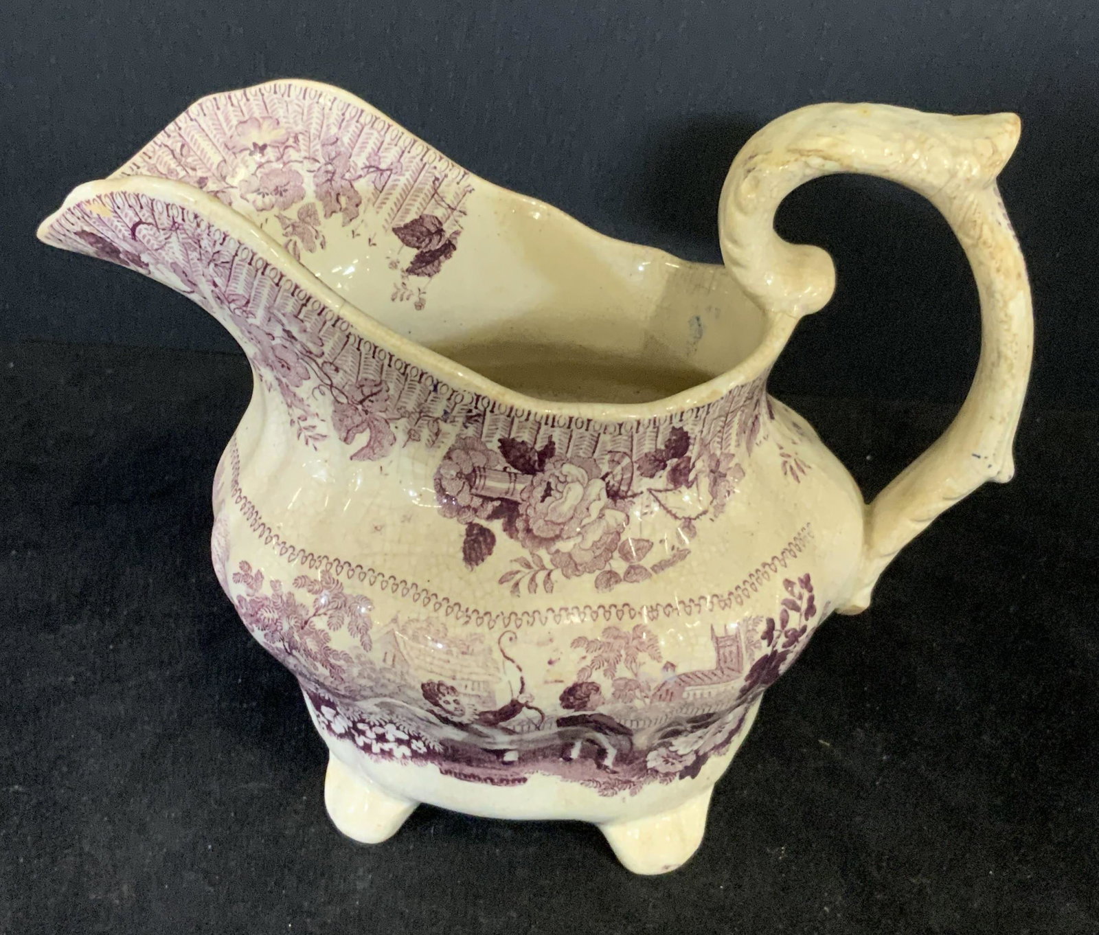 Trademarked Vintage Porcelain Footed Cream Pitcher: A vintage porcelain cream / cream pourer with purple designs. The designs feature people, foliage, and florals. The people are holding bows and conversing. One faces forward, the other faces away. Dec
