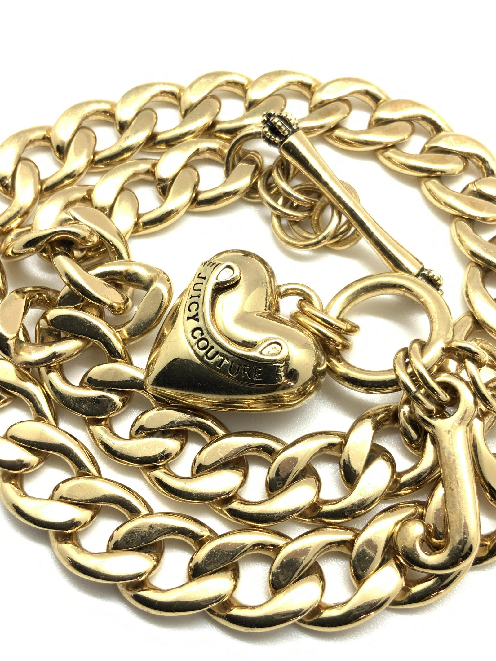 JUICY COUTURE Curb Chain Heart Necklace, Jewelry: A polished gold tone curb chain necklace. A heart bobble hangs near the bar clasp closure. The heart reads: JUICY COUTURE in etched metal. Next to the heart bobble is a letter ‘ J ‘ bobble. Neckla