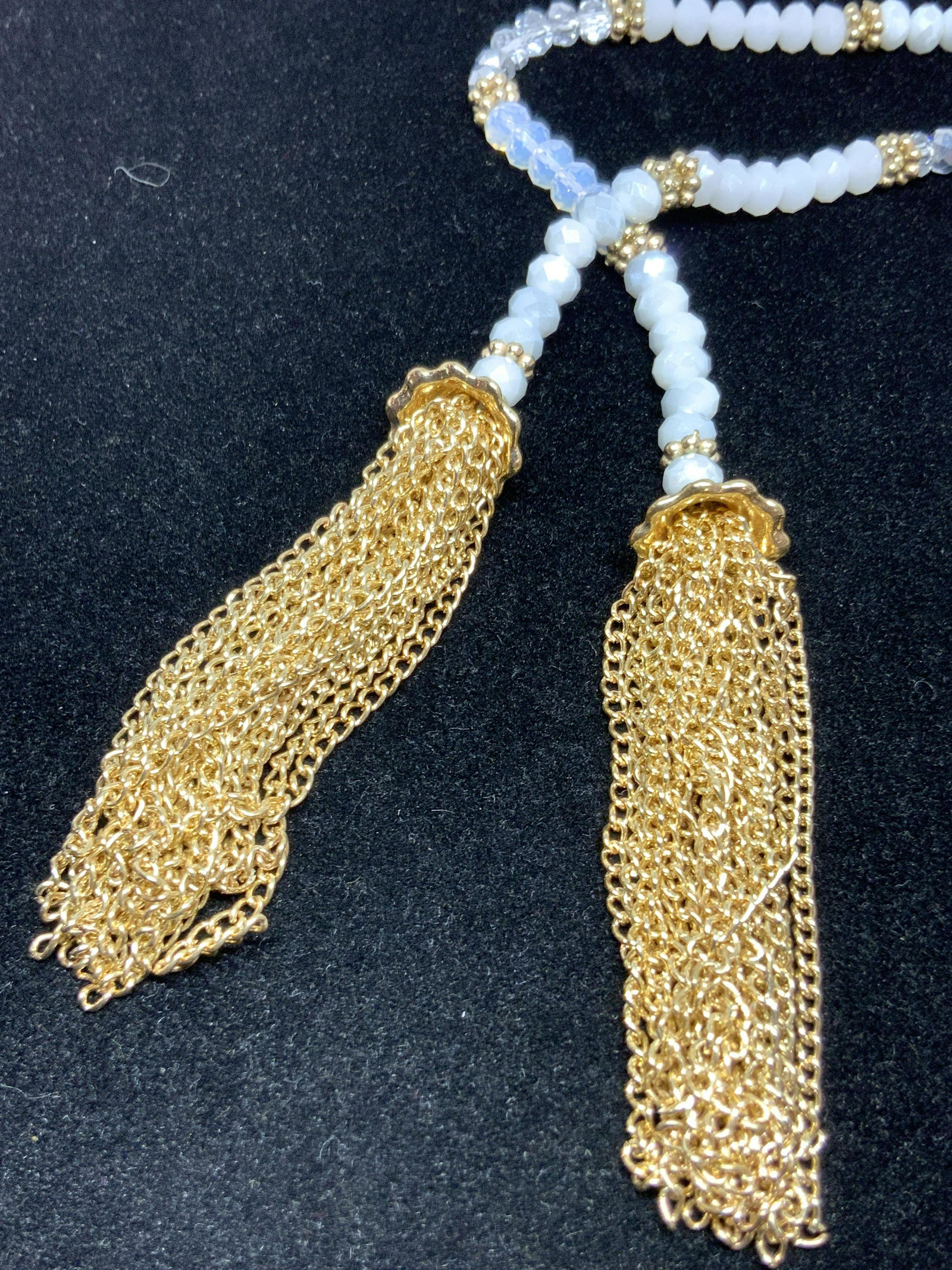 Iridescent Tassel Tipped Beaded Lariat Necklace: A beaded lariat necklace covered in faceted spherical beads. Beads have an iridescent coating and are colored in various hues such as silver tone metallic, milky white, colorless, and various hues in 