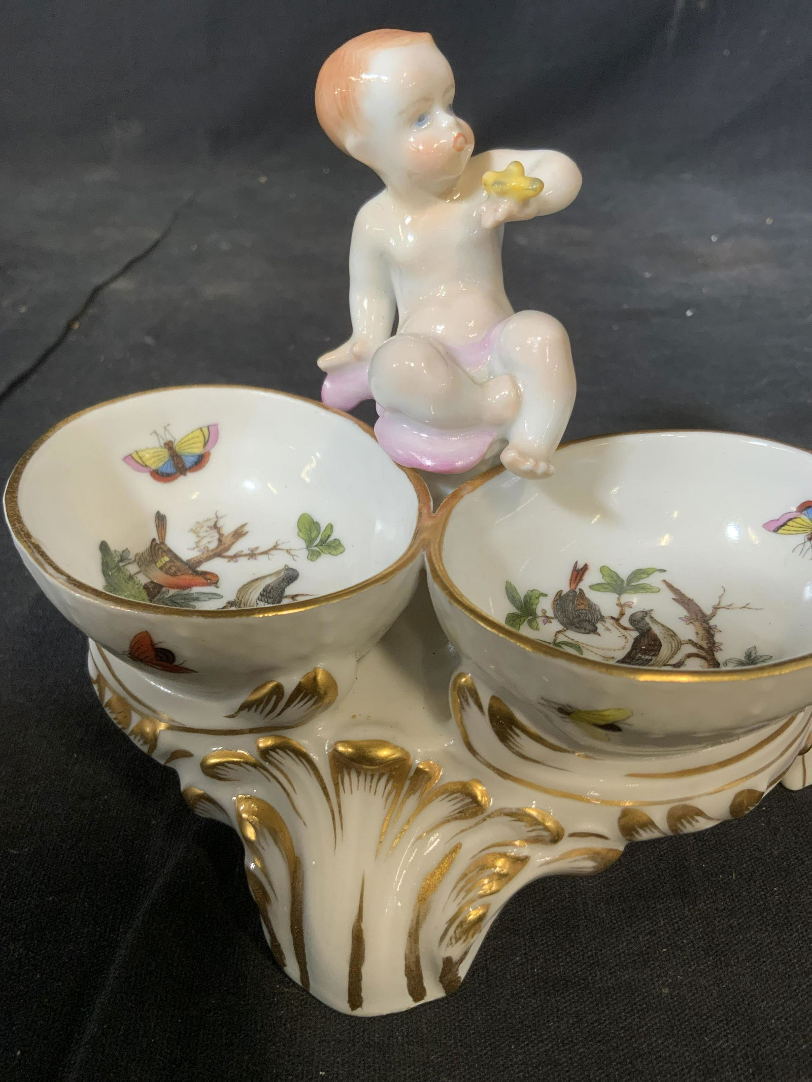 HEREND porcelain Putti Nut Dish: Marked for here d, Hungary and hall marked on underside, table accessory, design of putti figural with two bowls at base, with painted birds and butterflies, all sit on scrolled gilded base, decorativ