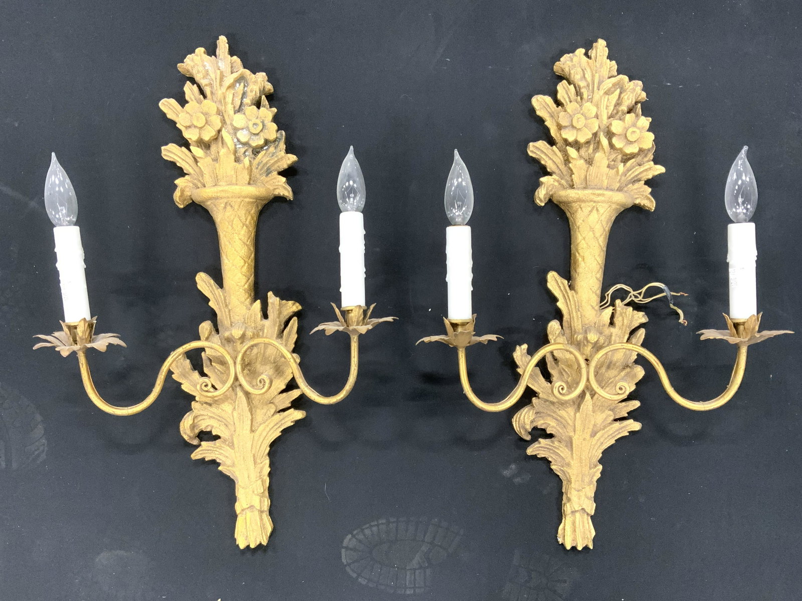 Pair 2 Arm Ornate Floral Detailed Wall Sconces: Wall sconces are gold toned and appears to be wooden. Arms have s curve detailed arms. Tops of sconces have floral & leaf detail. Each have candlestick style bulb sockets. Sconces, wall sconces, ornat