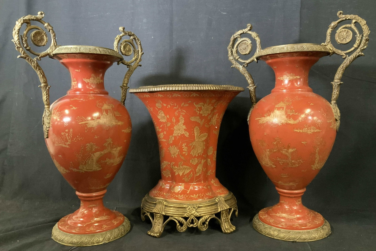 3 Pcs Chinese Porcelain Garniture Set: Three piece set includes two scrolled handled urns, centerpiece vase, each with gold toned metal, bases and trim, gold Asian figurative and garden designs on saffron toned base colors, each urn measur