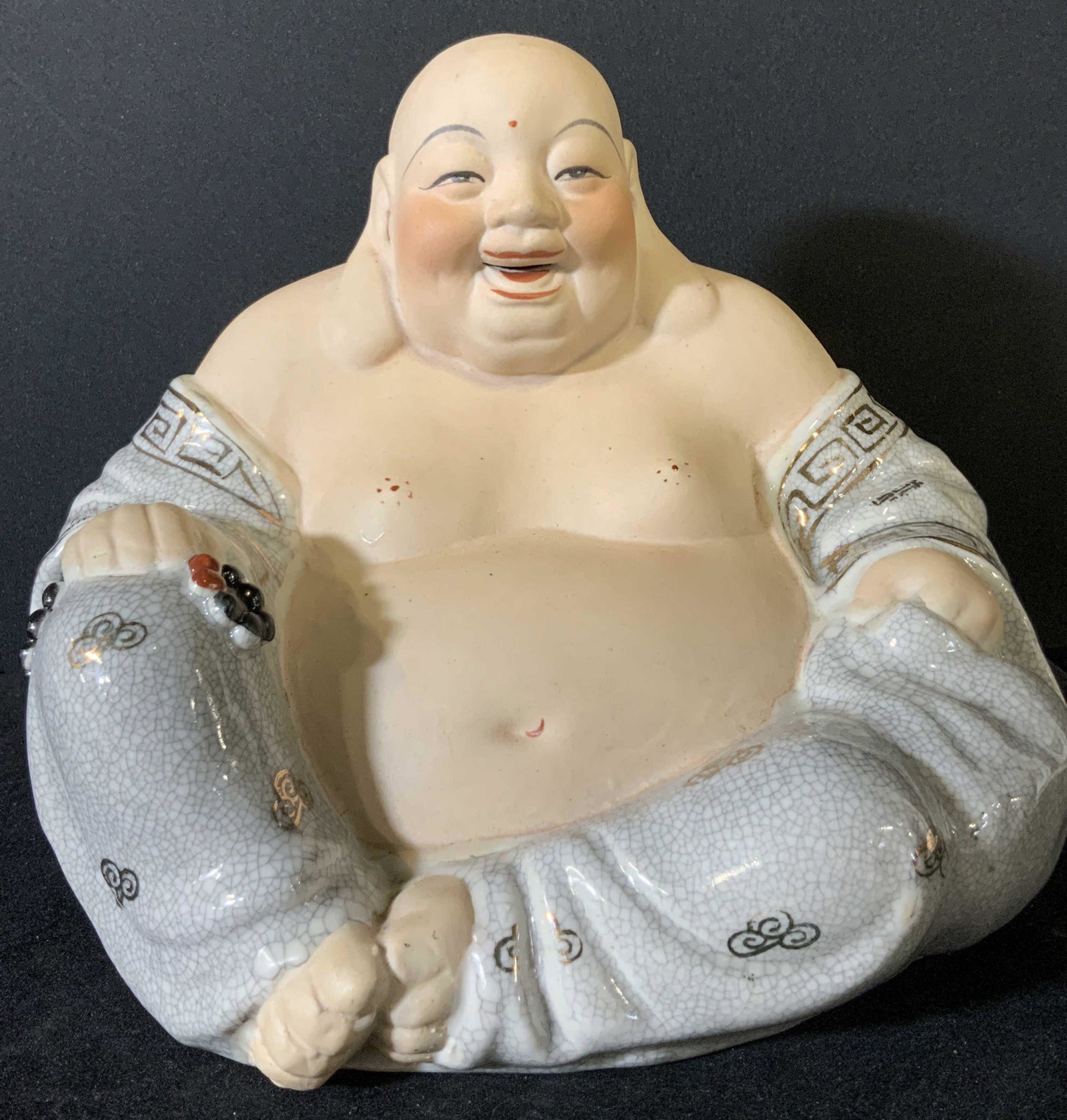 Ceramic Seated & Laughing Buddha Statue: A jovial Buddha depicted in a seated position and laughing. In his hands he holds embossed porcelain prayer beads. The robes and beads accenting Buddha are glazed and hand painted with gilt designs. B