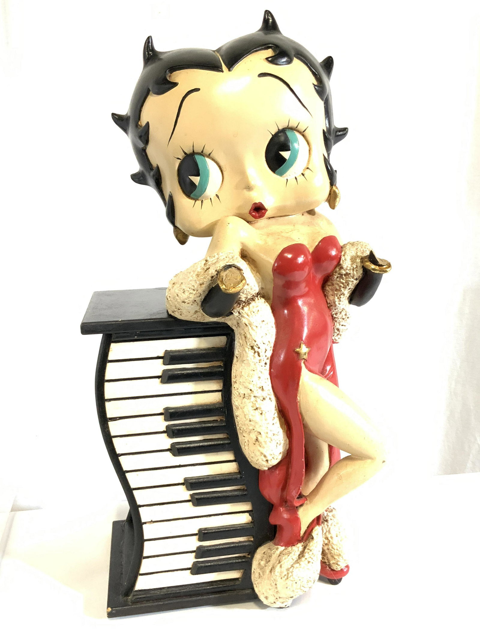 HEARST Composite Floor Sized BETTY BOOP Statue: A composite statue depicting Betty Boop leaning against a piano. Betty Boop is depicted with her classic hair style and red dress. There are metallic gold tone accents; her earrings, cuffs, and the st