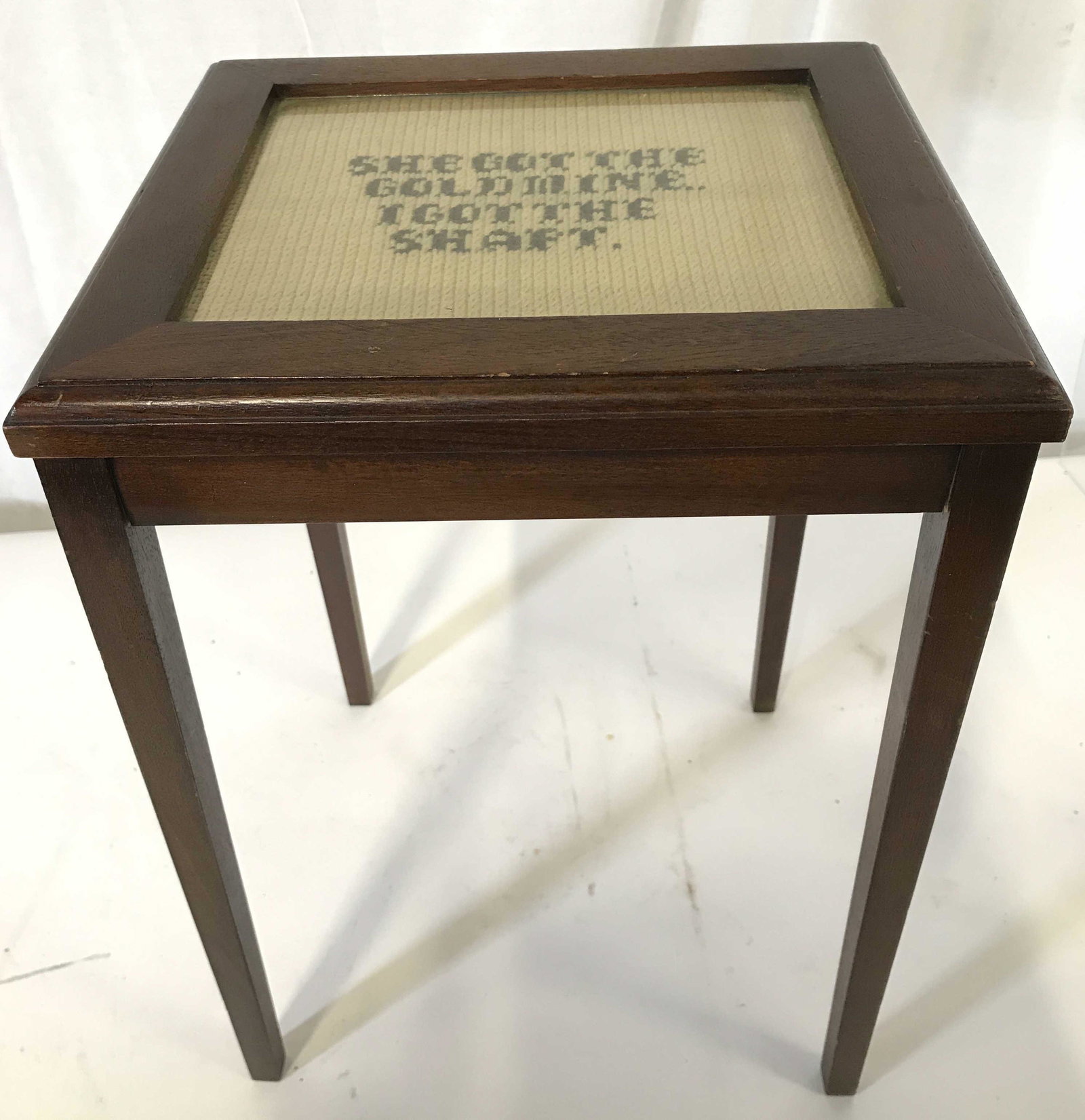 Vintage Side Table W Needlepoint & Glass Top: Side table is wooden and vintage. Piece has needlepoint detail and glass top. Needlepoint detail reads, SHE GOT THE GOLDMINE I GOT THE SHAFT. Piece measures approximately 17 inches tall 12 inches wide