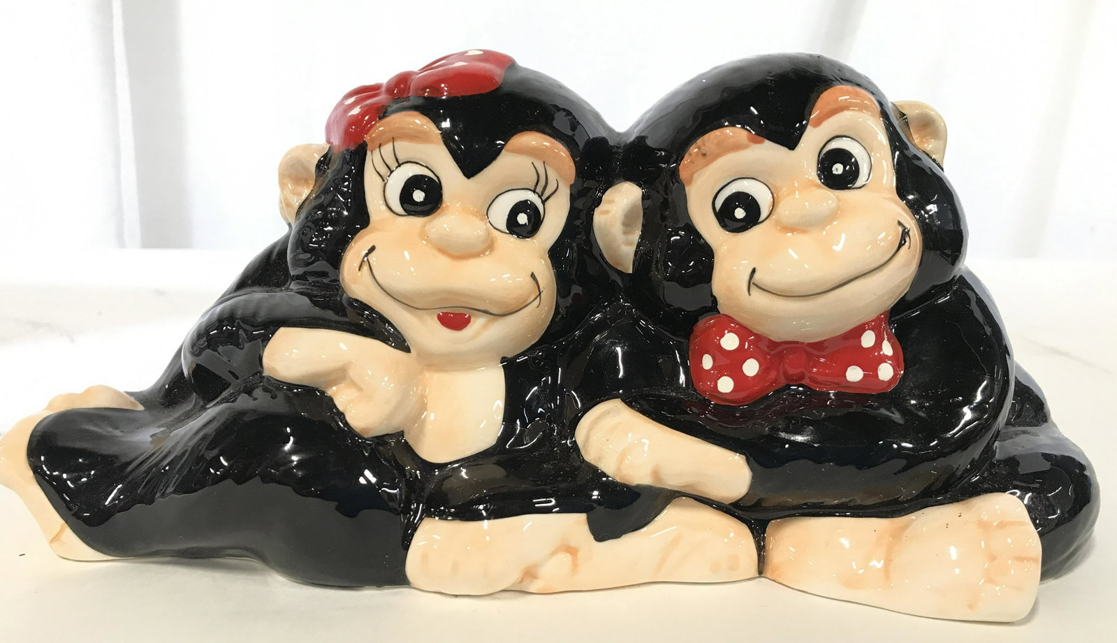 Porcelain Monkey Figural Piggy Bank: Piece is porcelain. Depict a male and female monkey. One has red and white polka dotted bow tie the other has red and white polka dotted head bow. Piece has 2 coin slots on backs of head areas. Piece 