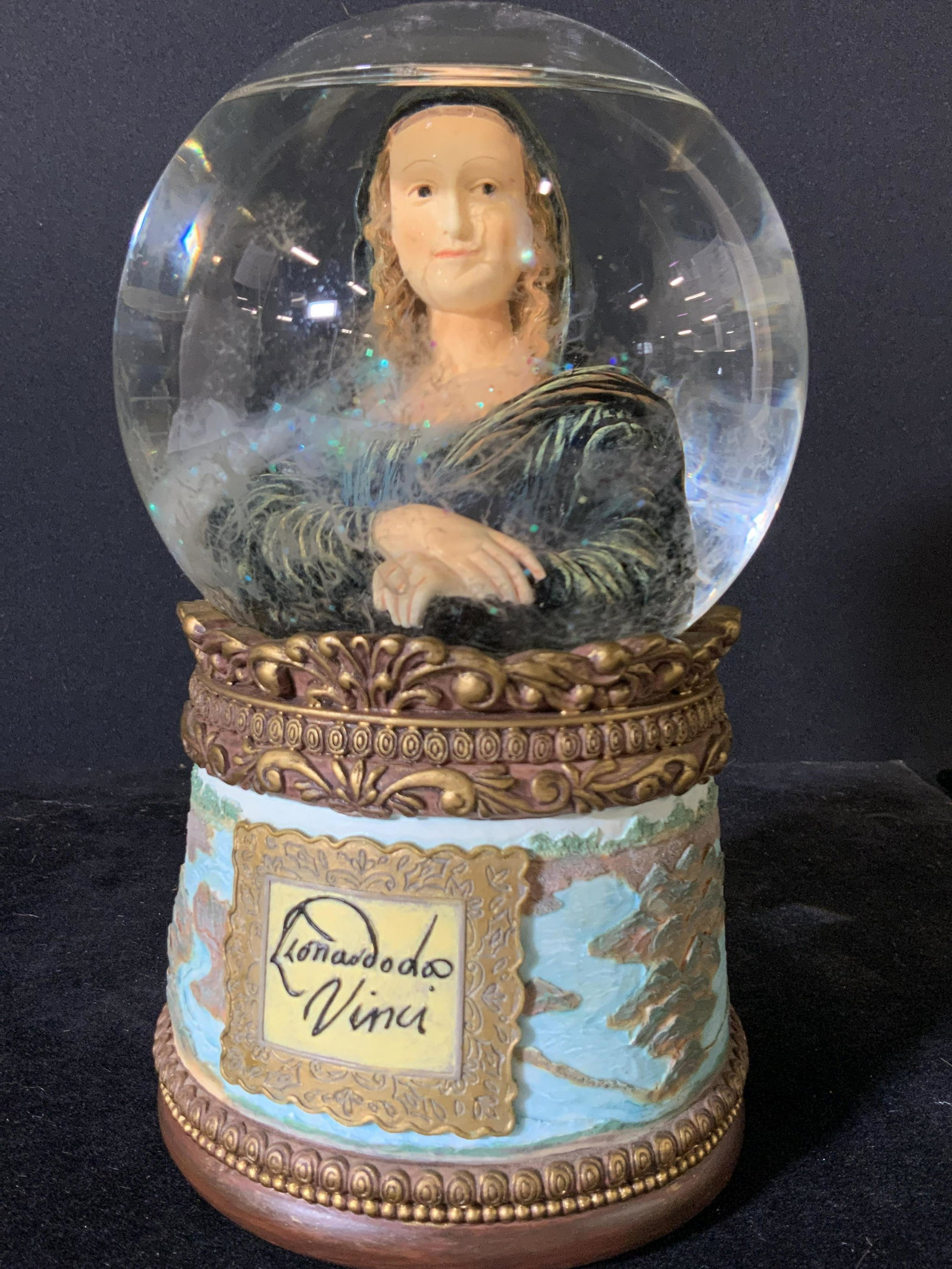 Art Glass MONA LISA Tabletop Snow Globe: A snow globe comprised of ceramic and glass. Etched ceramic designs all along the base of the snow globe. The frontside reads: Leonardo Da Vinci. The backside reads: Mona Lisa 1503-1506. The glass of 