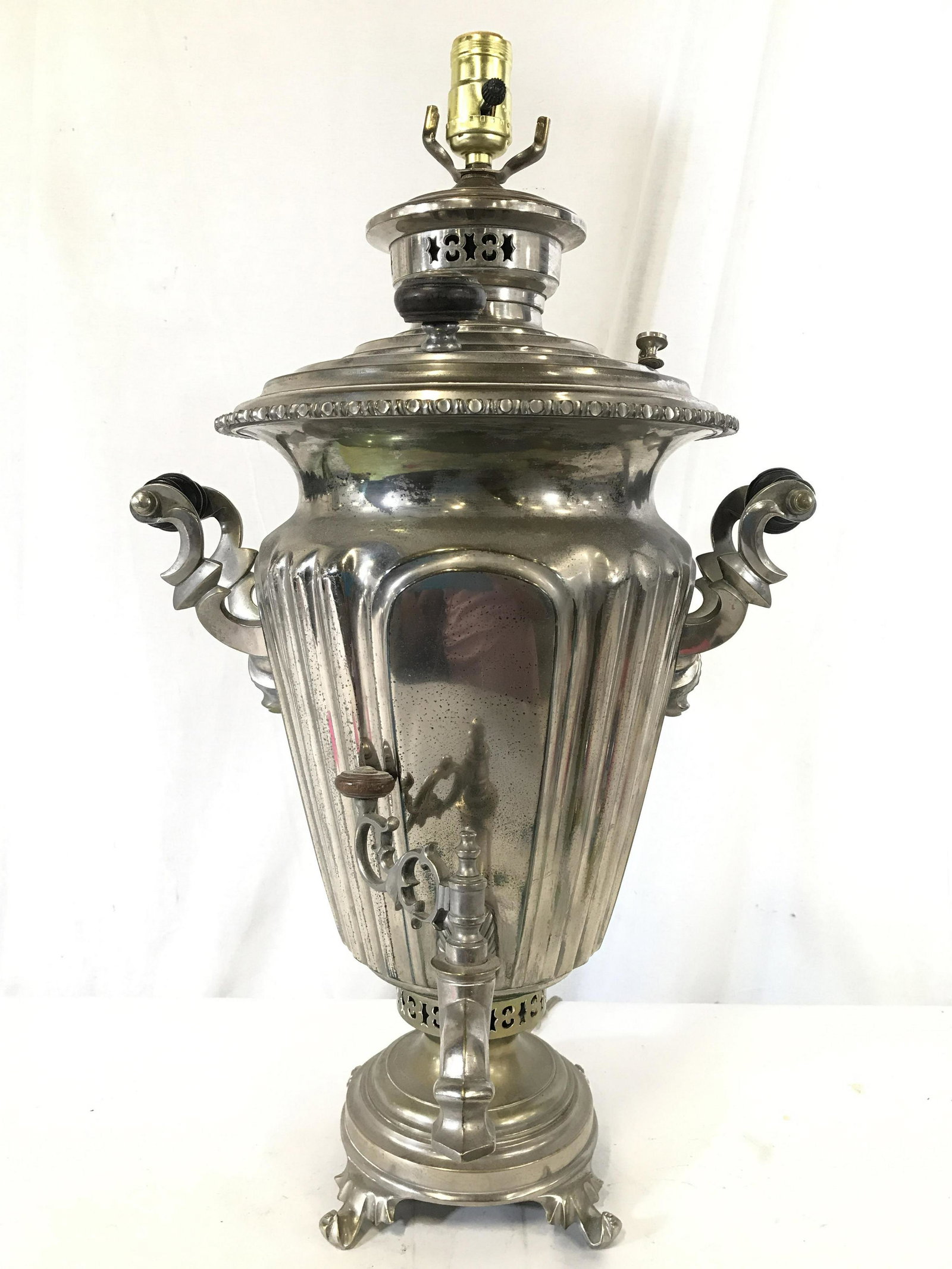 Antique Russian Samovar Lamp W Handles: Samovar is silver toned and metal. Piece has 2 black toned handles. Base is pedestaled with 4 feet. Piece has twisting spigot handle. Samovar has pierced geometric detail near top & base. Piece measur