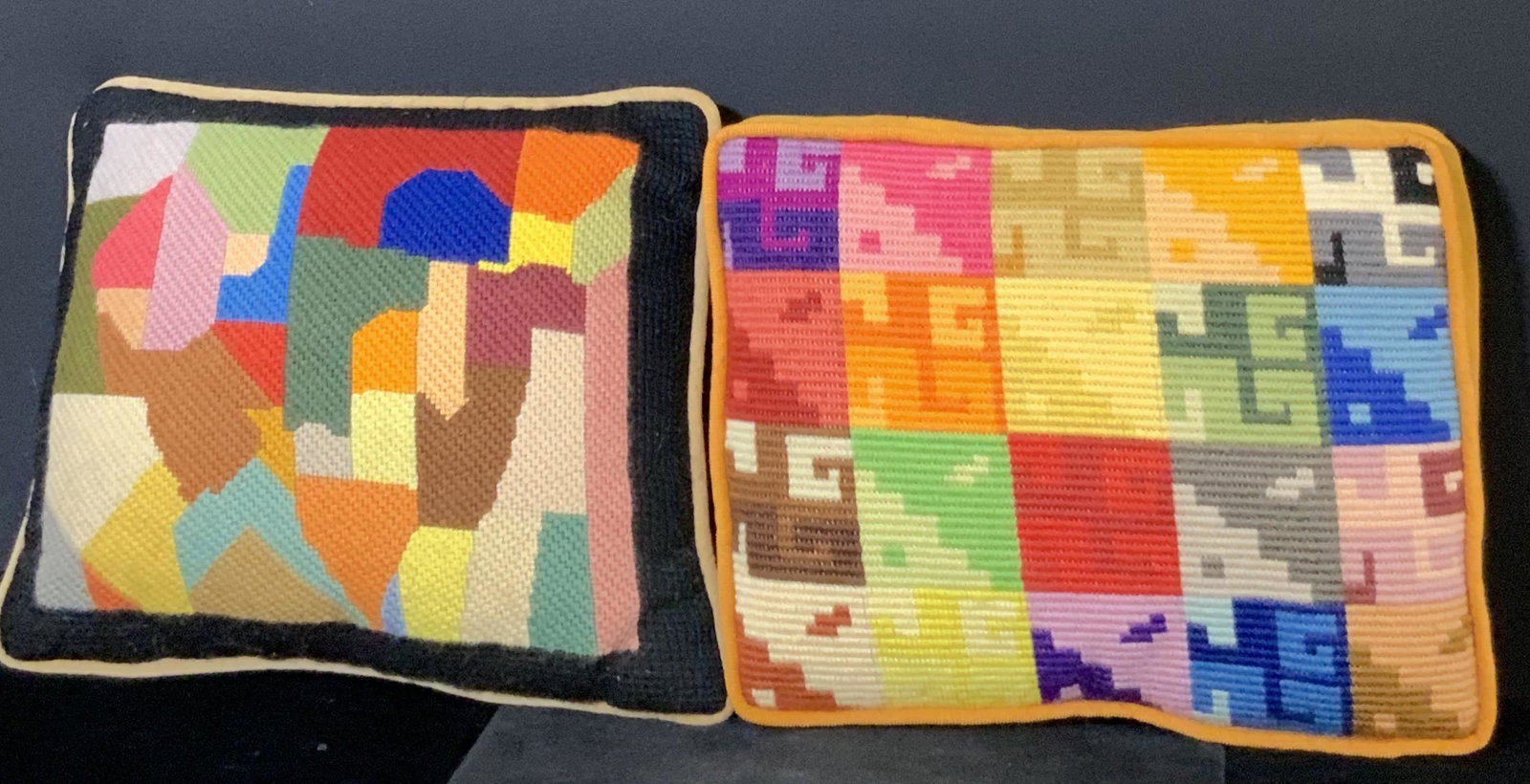 Lot 2 Abstract Needlepoint Throw Pillows: Two colorful, abstract needlepoint throw pillows. Vibrant hues of reds, blues, yellows and more adorn these pillows. The underside of the needlepoint pillow with black boarding is a velour material. T