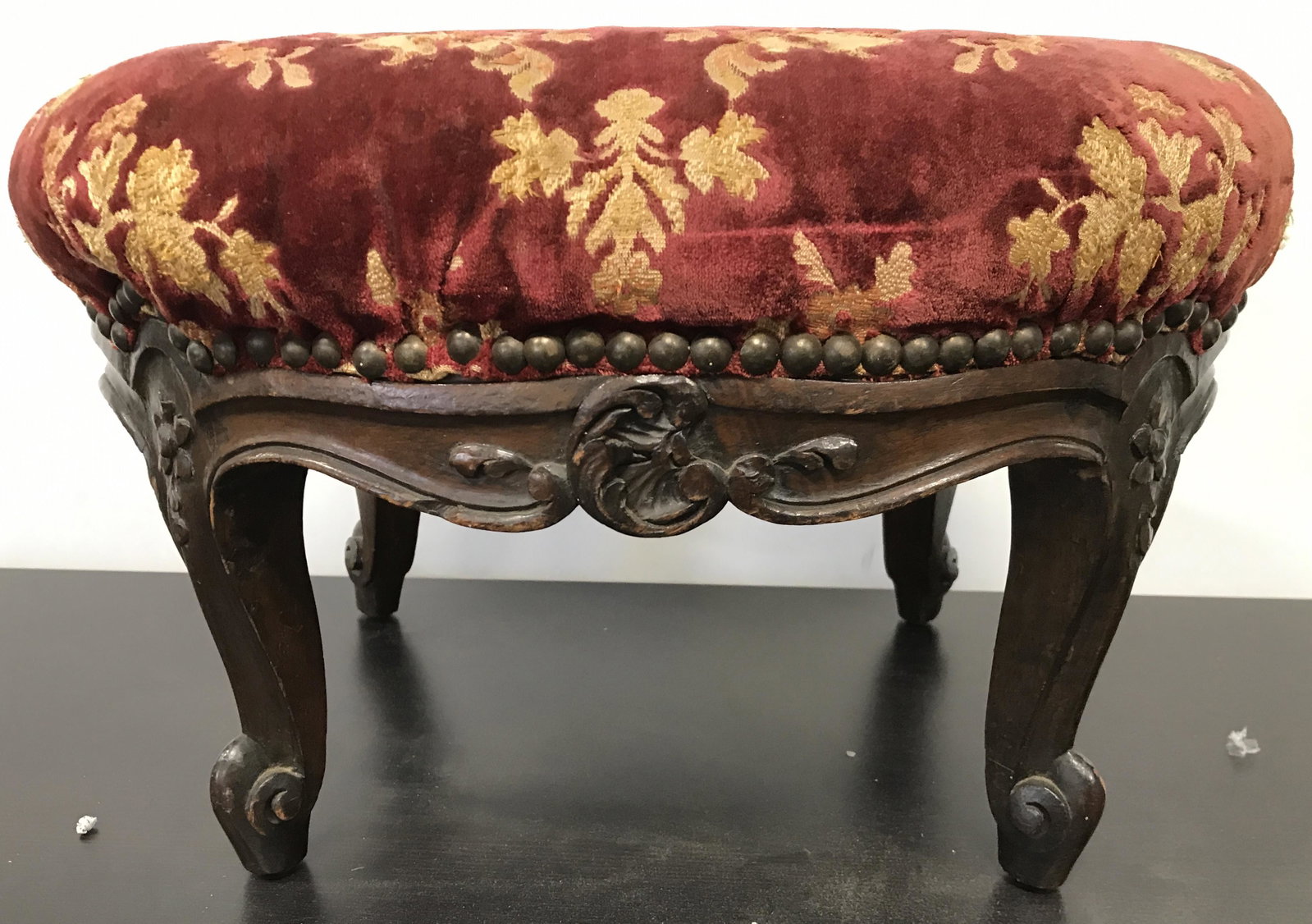 Antique Carved Wooden Upholstered Footstool: Footstool is antique. Piece has red & gold toned floral leaf and vine detail. Edge of upholstery has nailhead detail. Legs and feet appear to possibly be hand carved. Piece measures approximately 16 i