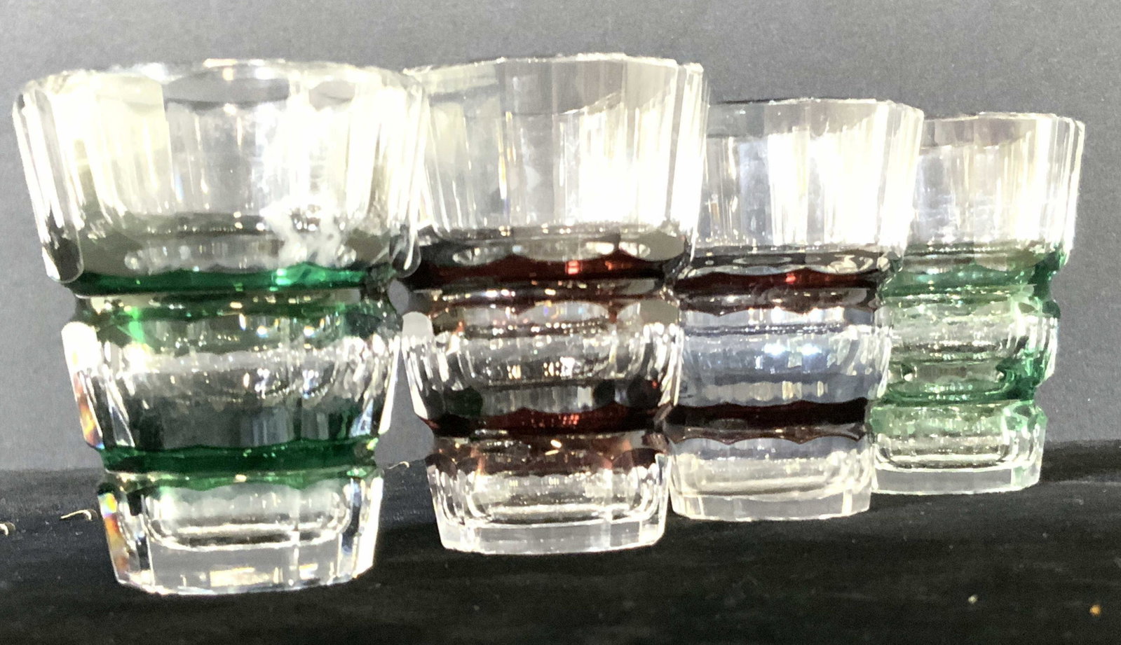 Set 4 Cut Crystal Green & Maroon Shot Glasses: Four cut crystal art glass shot glasses. Two feature green accents, two feature maroon accents. Approx. 3H x 2W in. One glass is slightly shorter than the others. Mild discoloration on interior of cry