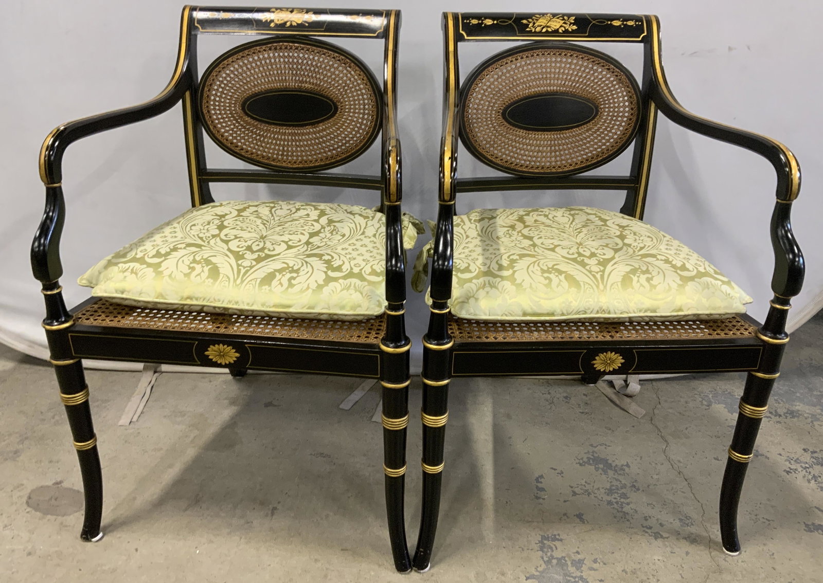 Pair Vintage Caned Regency Style Armchairs: Pair of vintage wooden armchairs. Each chair is hand painted black with gilt highlights and detailing. Backs and seats are crafted fro, woven cane. The top of each chair back is decorated with illustr