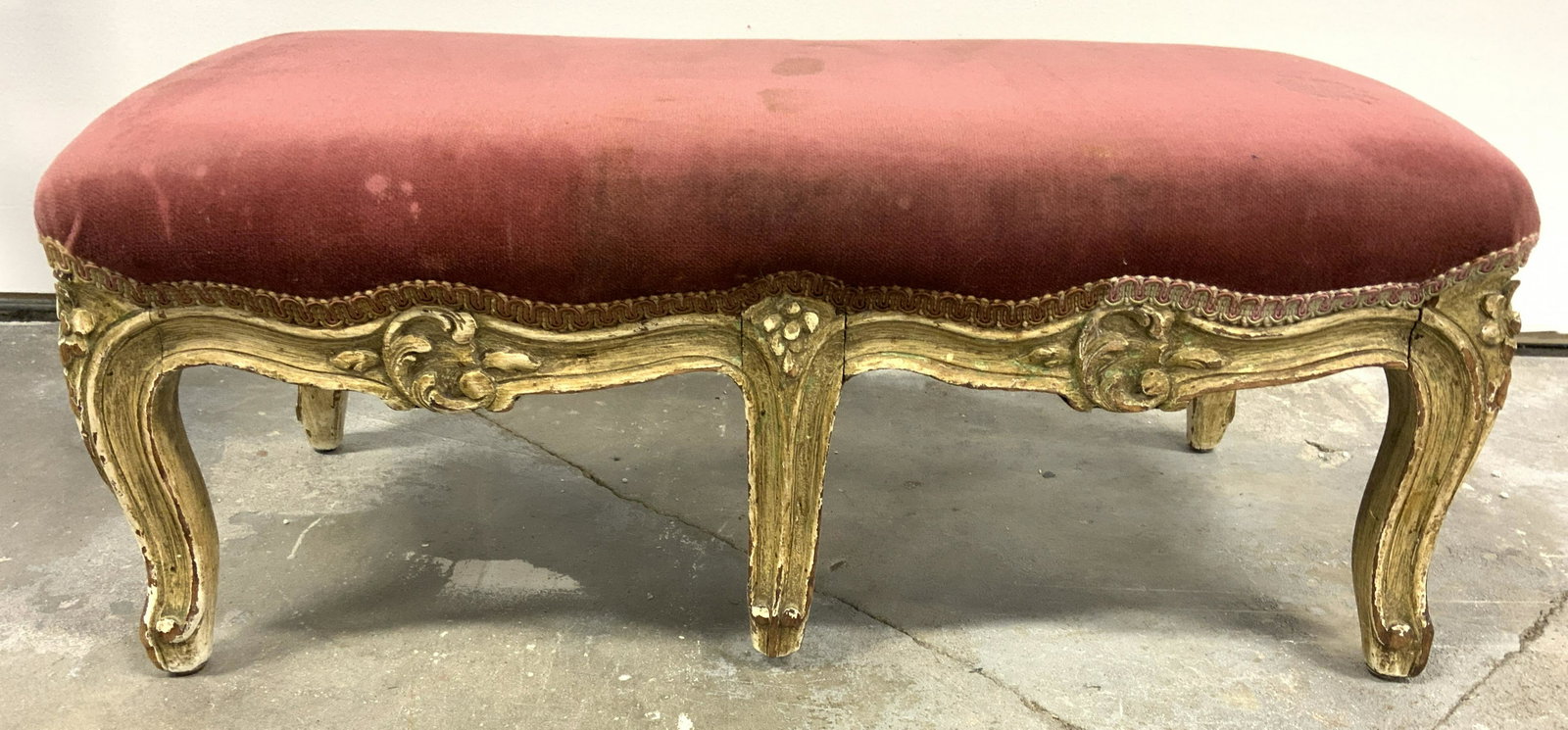 Antique Carved Wooden Upholstered Footstool: Footstool is antique. Piece has 6 carved wooden legs & feet. Upholstery appears to possibly be crushed silk. Wooden area has carved floral & c scroll detail. Piece measures approx 23 inches long 12 in