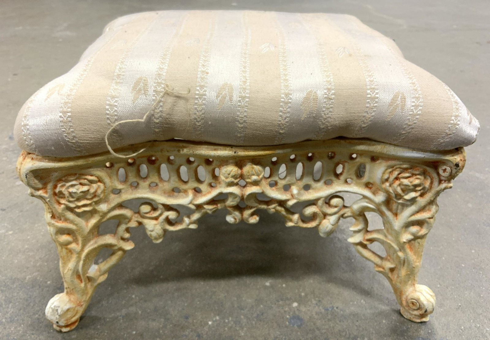 Vintage Upholstered Metal Base Footstool: Footstool is vintage possibly antique. Piece has intricate metal base with c scroll style feet. Footstool is upholstered with cream toned upholstery. Piece measures approx 11 inches wide and deep 8 in