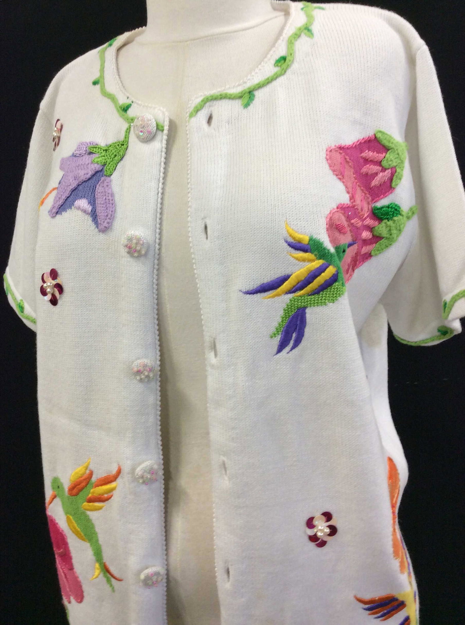 Quacker Factory Short Slv. Hummingbird Sweater: White knit sweater with many Textiles, faux pearls, sequins, embroidery, attractive hummingbirds suckling On flowers. Covered buttons. Size Med. 53% ramie 35 % Cotton 8% rayon 4 % acrylic. Couple spot