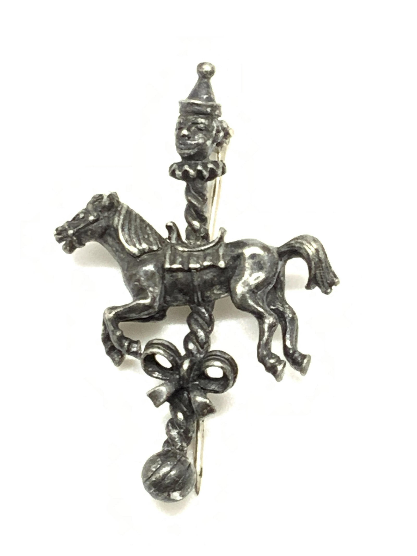 Vintage Clown Carousel Horse Brooch: A vintage deep gunmetal toned metal brooch. The piece features a clown face connected to a horse and a ribbon. The piece connecting them resembles a carousel pole. The connecting pole is twisted and t