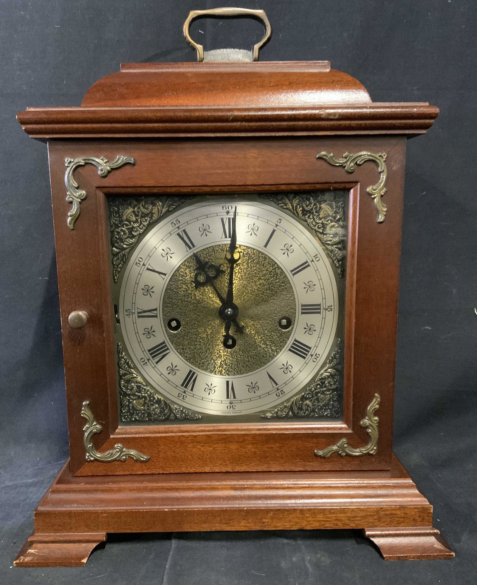 HAMILTON Wood Mantle Clock, Germany: Wooden HAMILTON mantle clock. Door on front opens, gives acces to clock face. Brass detailing decorates the clock. Clock face reads as HAMILTON, WESTERN GERMANY. A handle is mounted to the clock. Meas