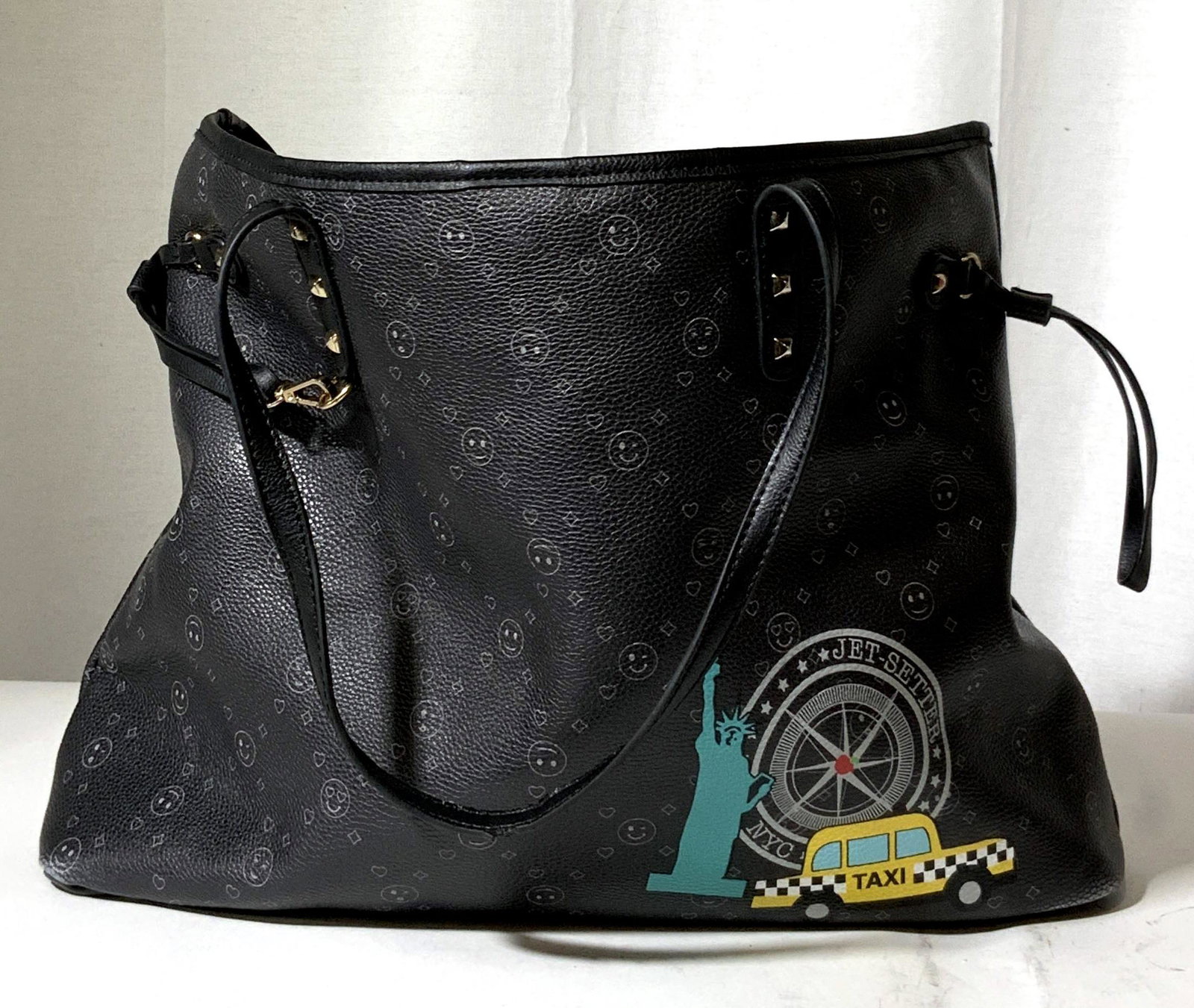 New York Taxi Tote Bag: Black tote bag with emoji designs throughout. Bottom right corner decorated with Statue of Liberty, compass, and taxi. Wording reads: NYC, Jet-Setter. Exterior is possibly patent leather, interior sof