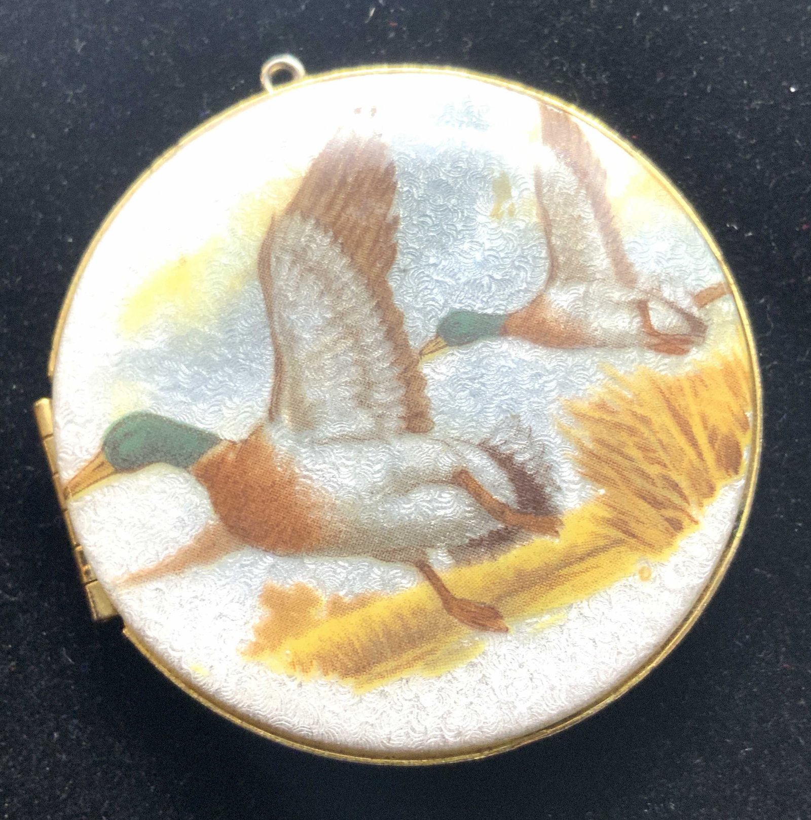 Hand Painted Enamel Duck Locket Pendant, Jewelry: A locket pendant crafted from gold tone metal and enamel. Hand painted scene depicts mallards / ducks taking flight. The background features blue skies, yellowing grass, and clouds. Piece opens with h