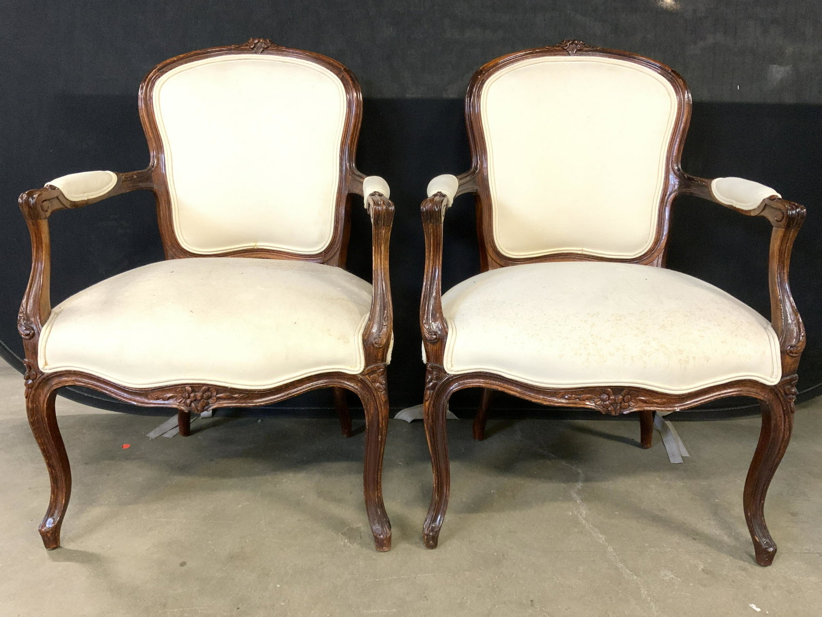 Pair Upholstered Carved Wooden End Chairs: Lot includes 2 end chairs. Each are wooden with carved floral detail. Each have cream toned upholstery with double welting at edges. Piece has partially upholstered armrests. Each measures approx 34 i