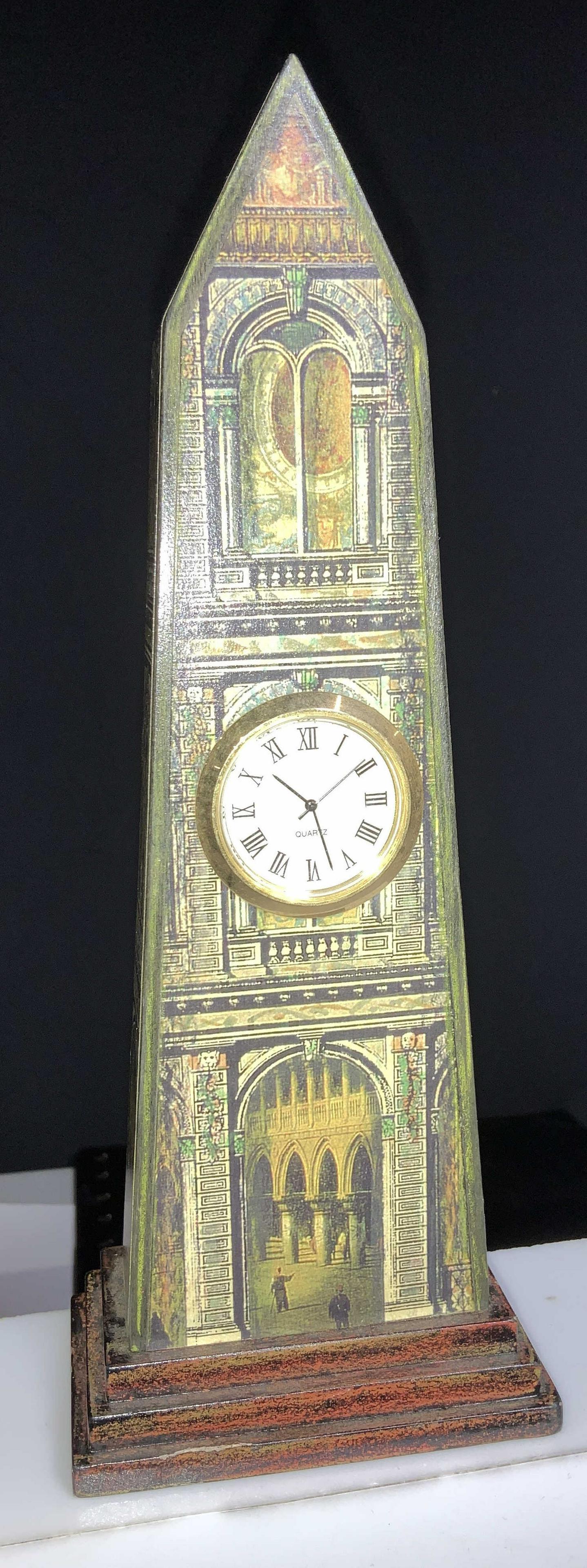 Signed Hut Studios NYC Obelisk Quartz Clock: Copyrighted hallmark signature , reads Hut Studios NYC. Obelisk with inserted round brass quartz an along clock. Decor depicts medieval structures of Venetian architecture. Light weight piece. 8.5 in