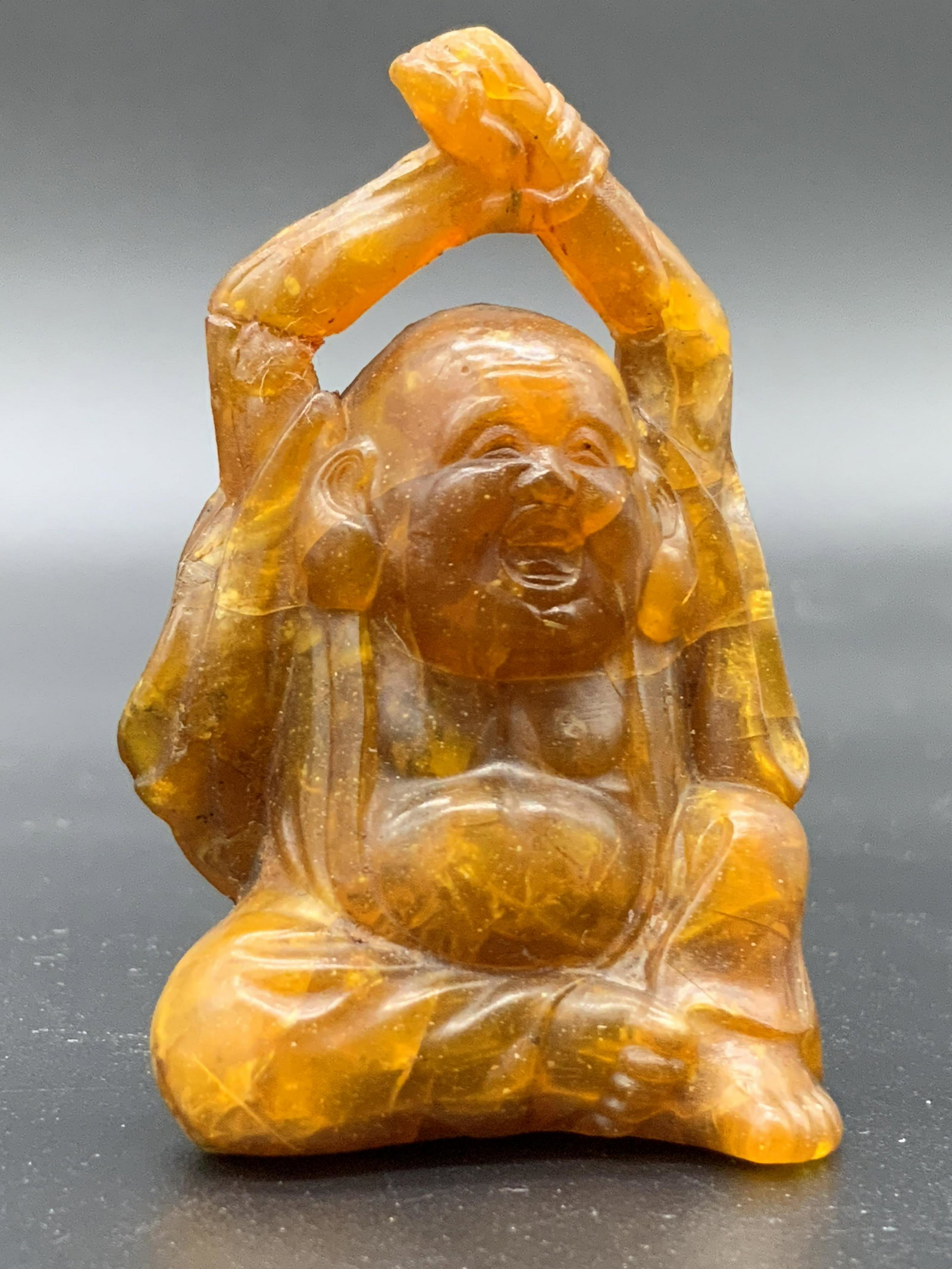 Vintage Collectible Carved Amber Buddha Figural: Vintage, possibly antique, collectible carved amber Buddha figural. The Buddha is depicted stretching and smiling. Two cord holes on the reverse. Mild surface cracks and cracks along the piece. Approx