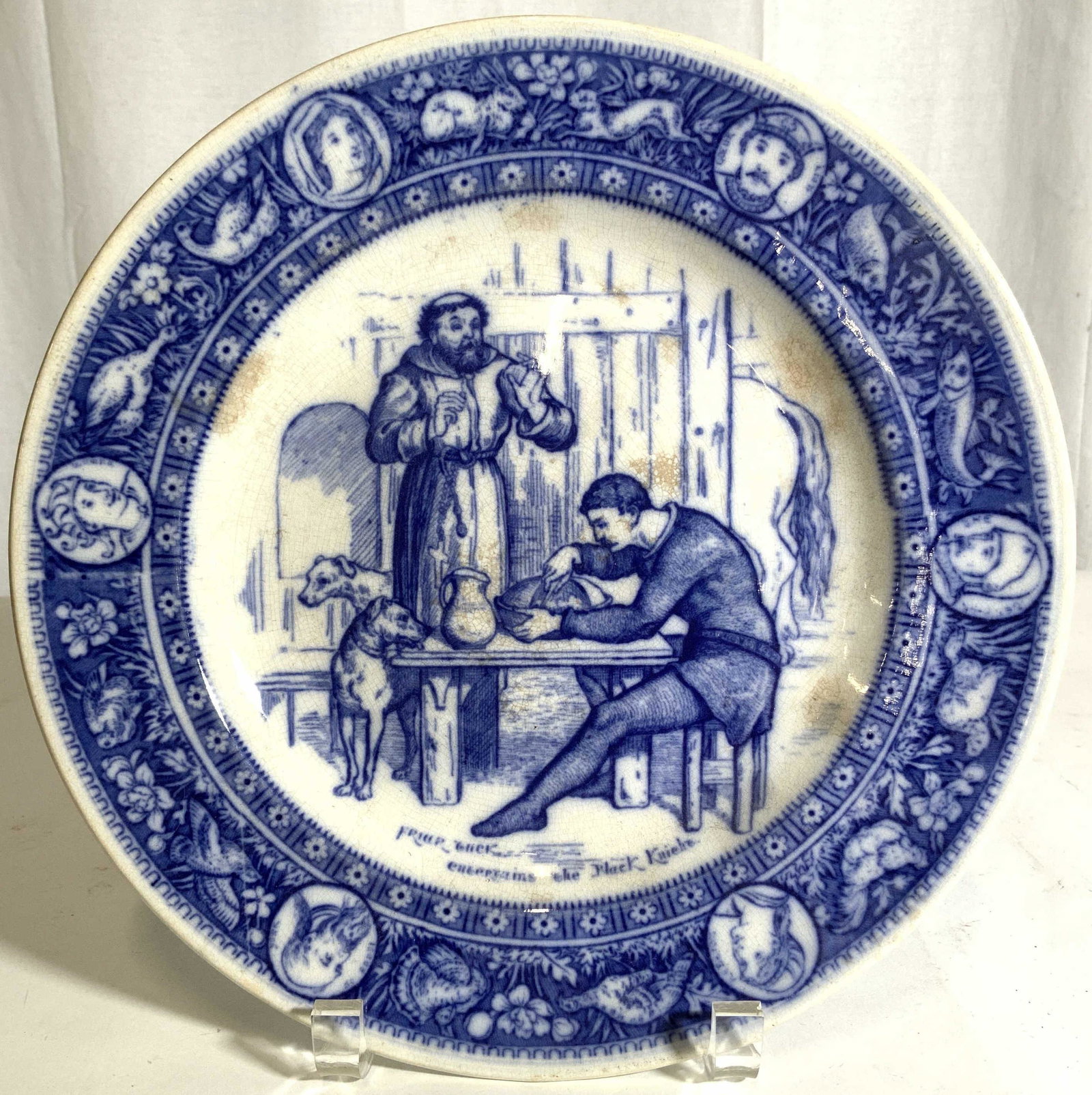 WEDGWOOD IVANHOE Porcelain Plate Chinaware: Underside is marked WEDGWOOD IVANHOE. Plate is white with intricate blue toned detail. Center has depiction of 2 dogs, one figure seated at a table, & 1 standing figure. Piece measures approx 10.5 inc