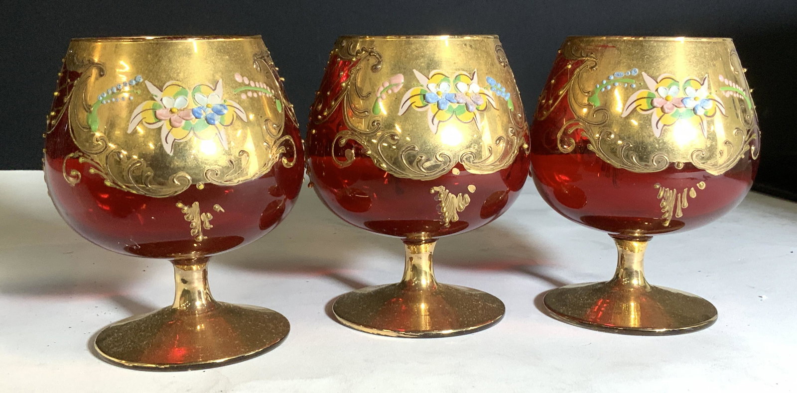Set 3 Gilt Cranberry Crystal Bohemia Goblets: These three goblets are exquisitely hand painted with heavy gilt cartouches, swirls and florals. Deep red color shines from exterior and interior. Stem all gilt,. No notice wear to paint. Vintage crys