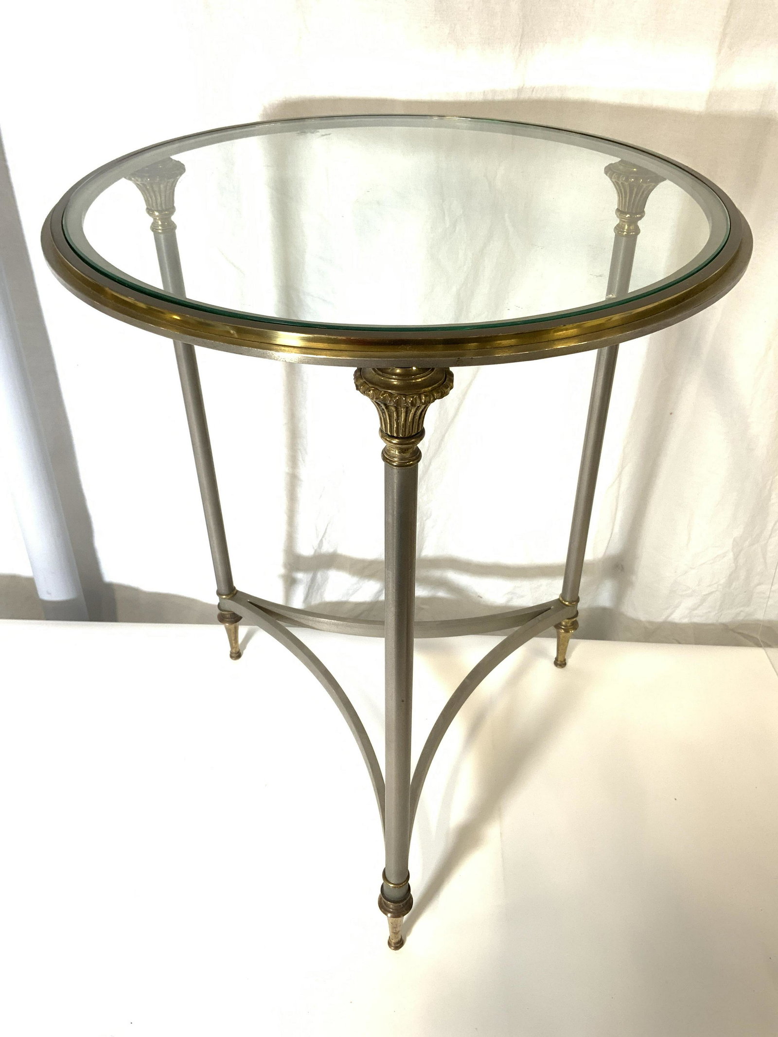 Vintage Brass & Metal Glass Top Round Side Table: Side table is vintage. Piece is brass and metal. Table top is round and has glass insert. Table has 3 gold toned brass feet. Table legs have curved stretchers in between . Piece measures approx 25 inc