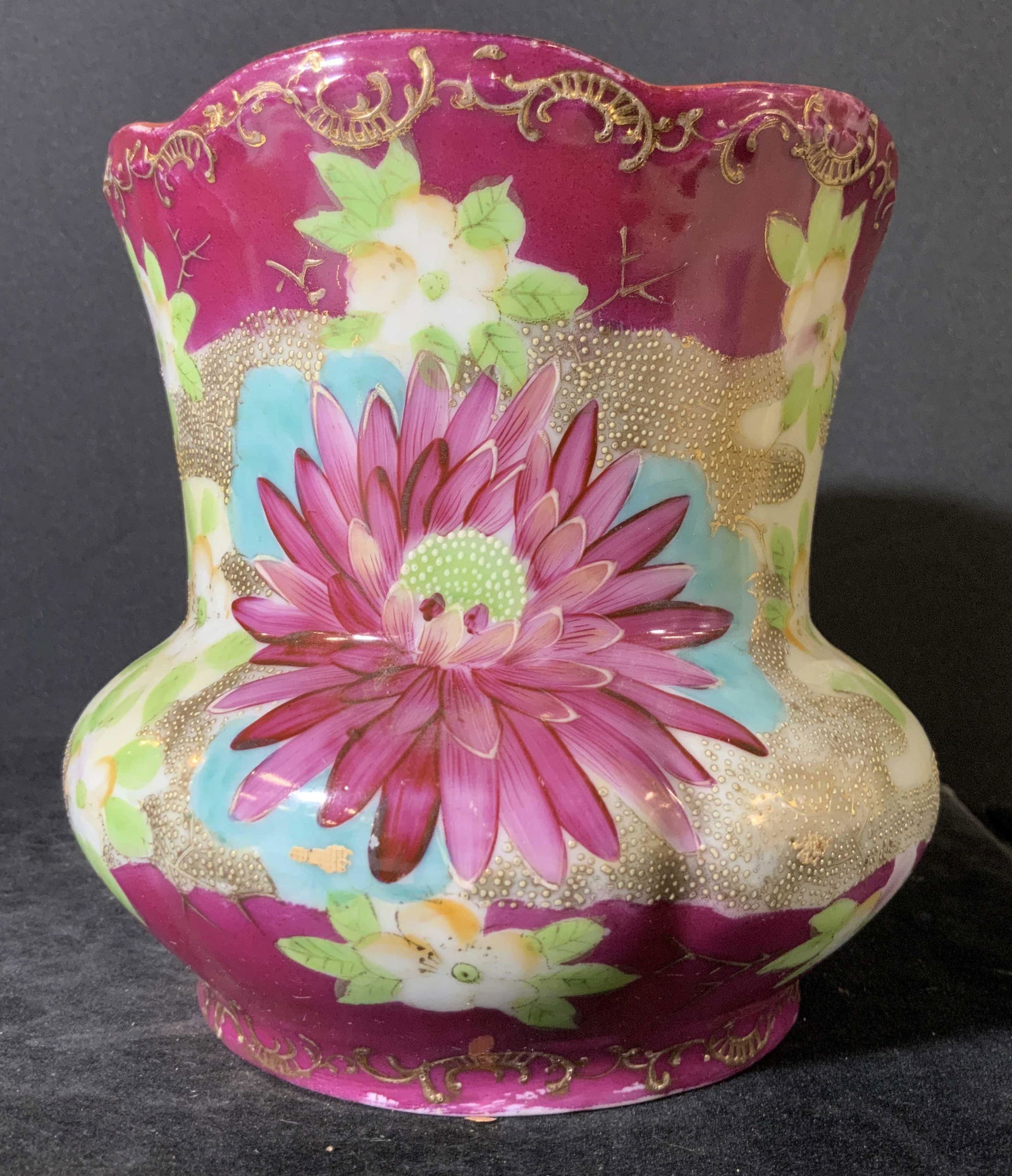 Hand Painted Floral Porcelain Tabletop Vase: A hand painted porcelain vase with a ruffled rim. The designs depict vibrant florals and gilt line work. Primarily purple toned with accents of green, yellow, and blue hues. Approx. 6H x 5W in. Some c