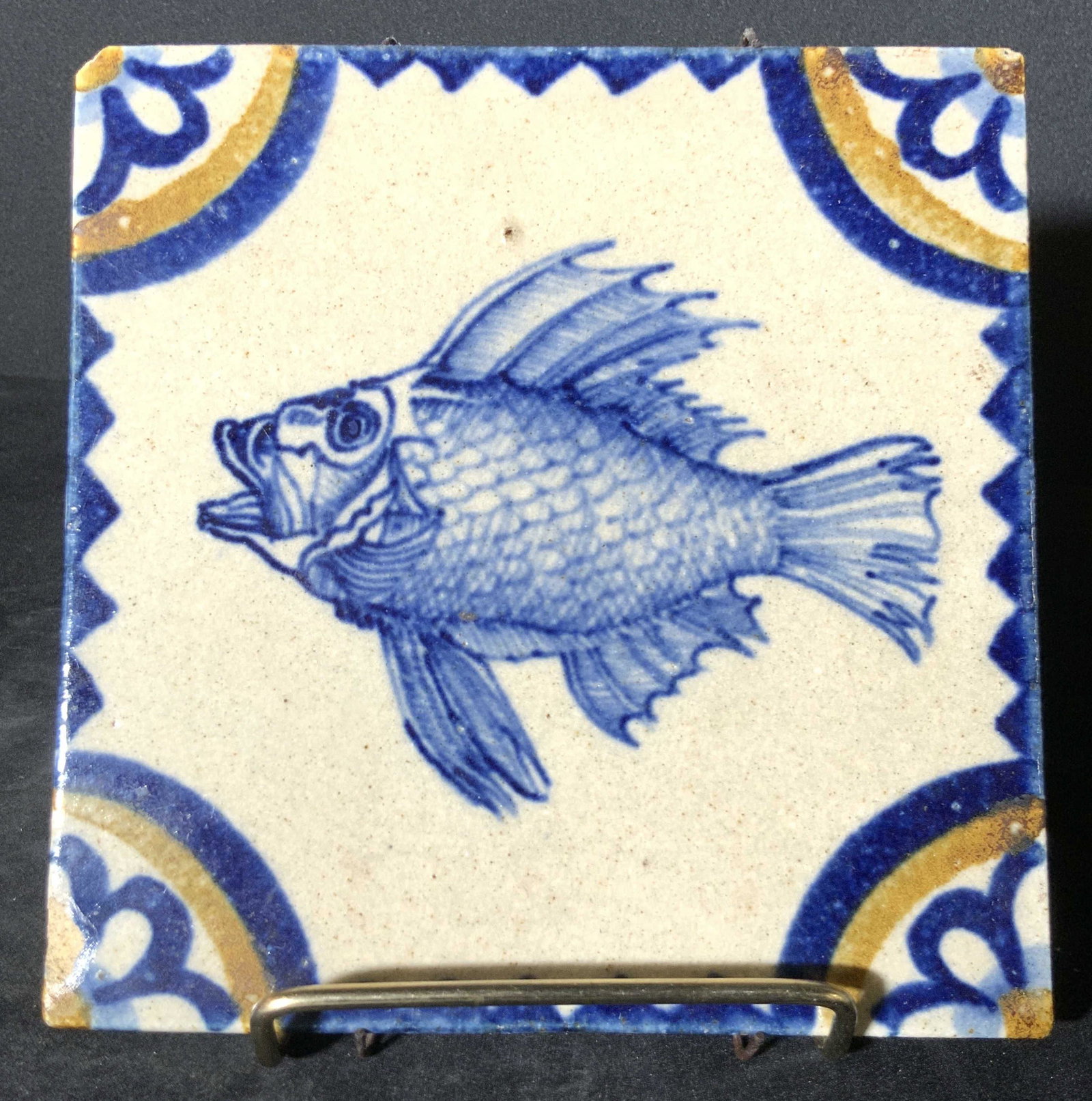 Vintage Glazed Ceramic Fish Motif Maritime Tile: Possibly vintage square glazed ceramic tile depicting a fish and florals in each corner. Painted blue and yellow hues designs. Underside has metal hardware for hanging. Some surface cracks, some mild 