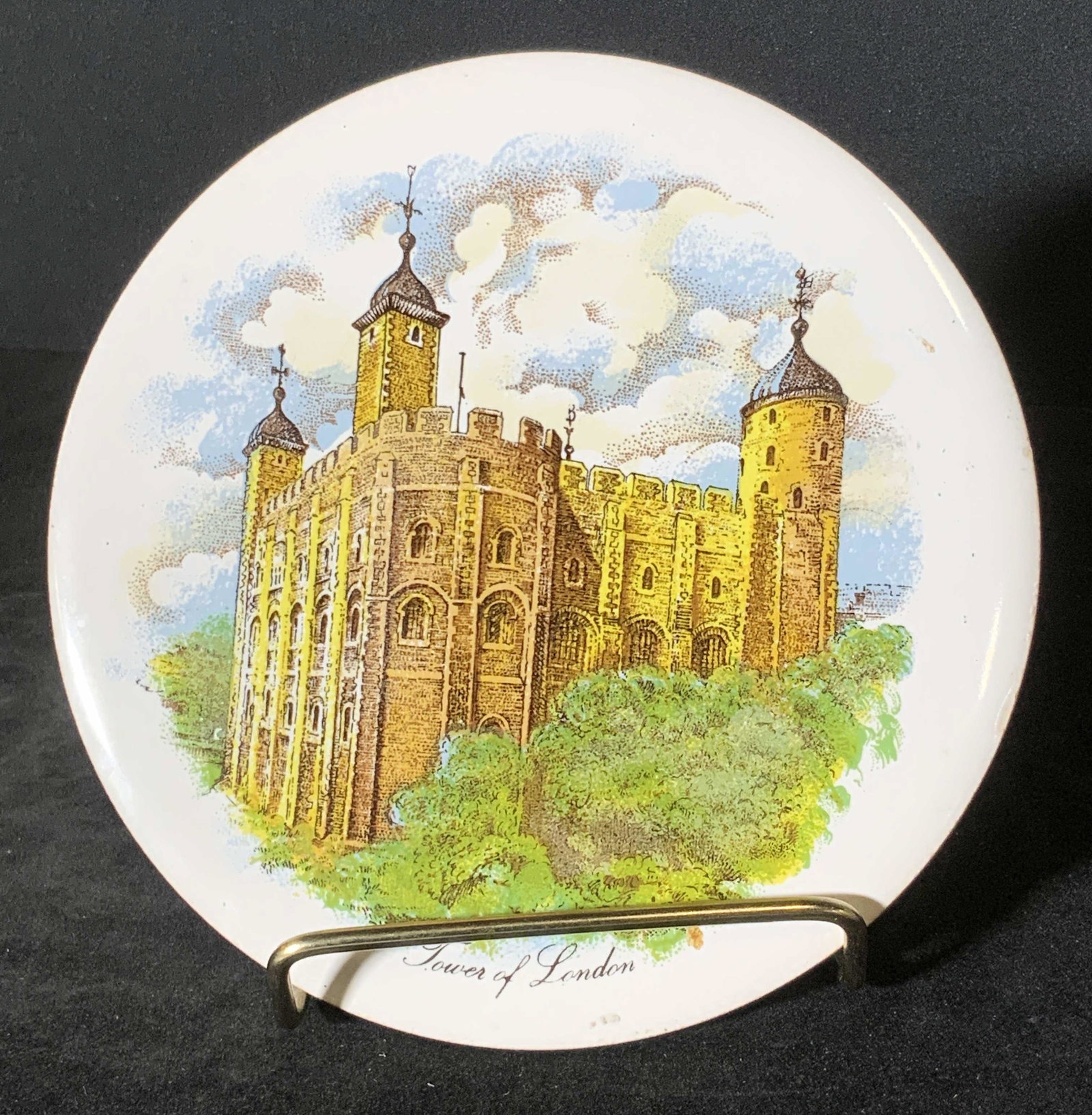 Signed British Ceramic Trivet, TOWER OF LONDON: A circular ceramic tile / trivet with three feet on the underside. The obverse depicts the Tower of London. Clouds decorate the background, trees decorate the foreground. Primarily yellow, white, blue