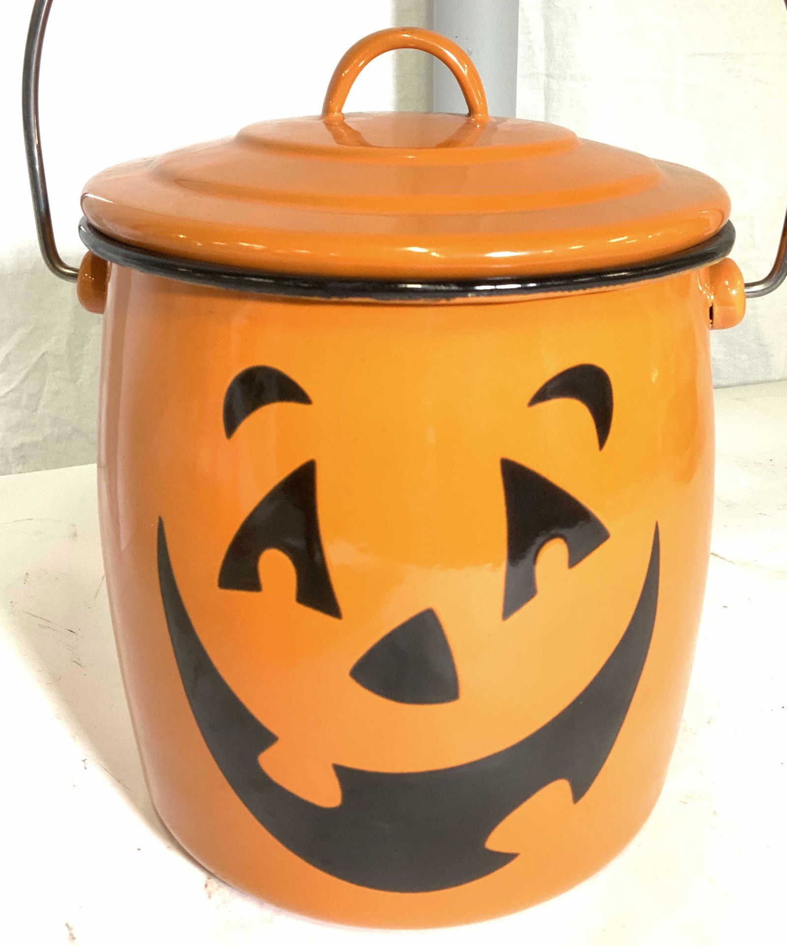 Orange Enamel Pumpkin Face Lidded Jar w/ Handle: Multi use enamelware, labeled HMK, pumpkin face, metal carry handle. Use as cookie jar, holding trick or treat candy, decorative object, cookware. Measures app 9 in ht. , 8 in width. Handle 9.5 in w.,