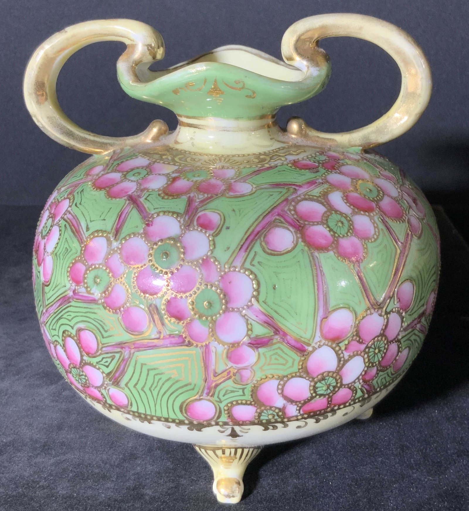 NIPPON Signed Asian Tabletop Porcelain Vessel: A pink and green hued floral vessel featuring in relief gold toned accents & gilt line work. The piece has a unique ruffled rim connected to twin handles. Four feet on the underside, signed NIPPON. so