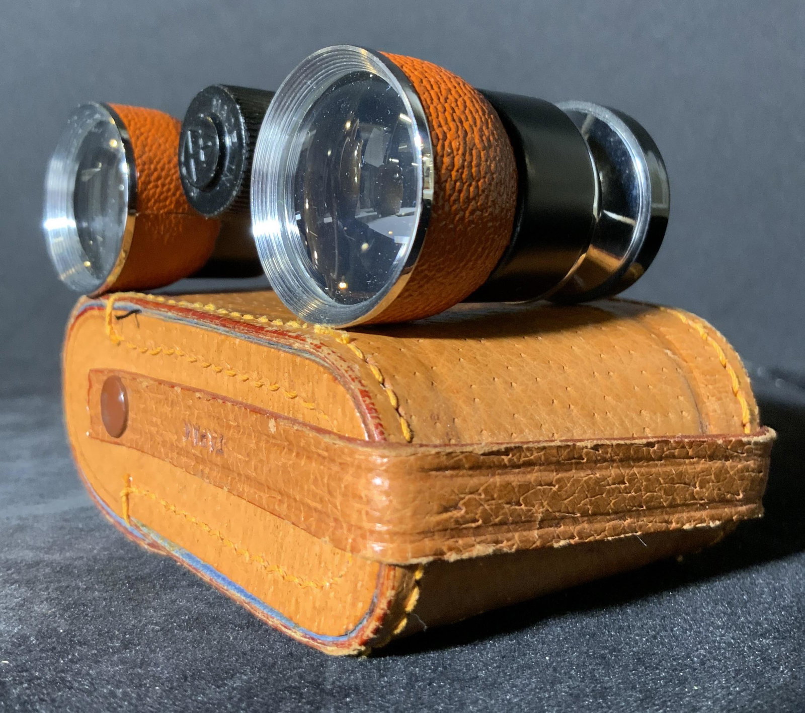 MILO Signed Leather Style Binoculars W. Case: Binoculars with light brown leather style accents and a light brown leather style case. The case snaps closed with metal hardware. Side of case reads JAPAN. The binoculars are crafted from metal. Silv