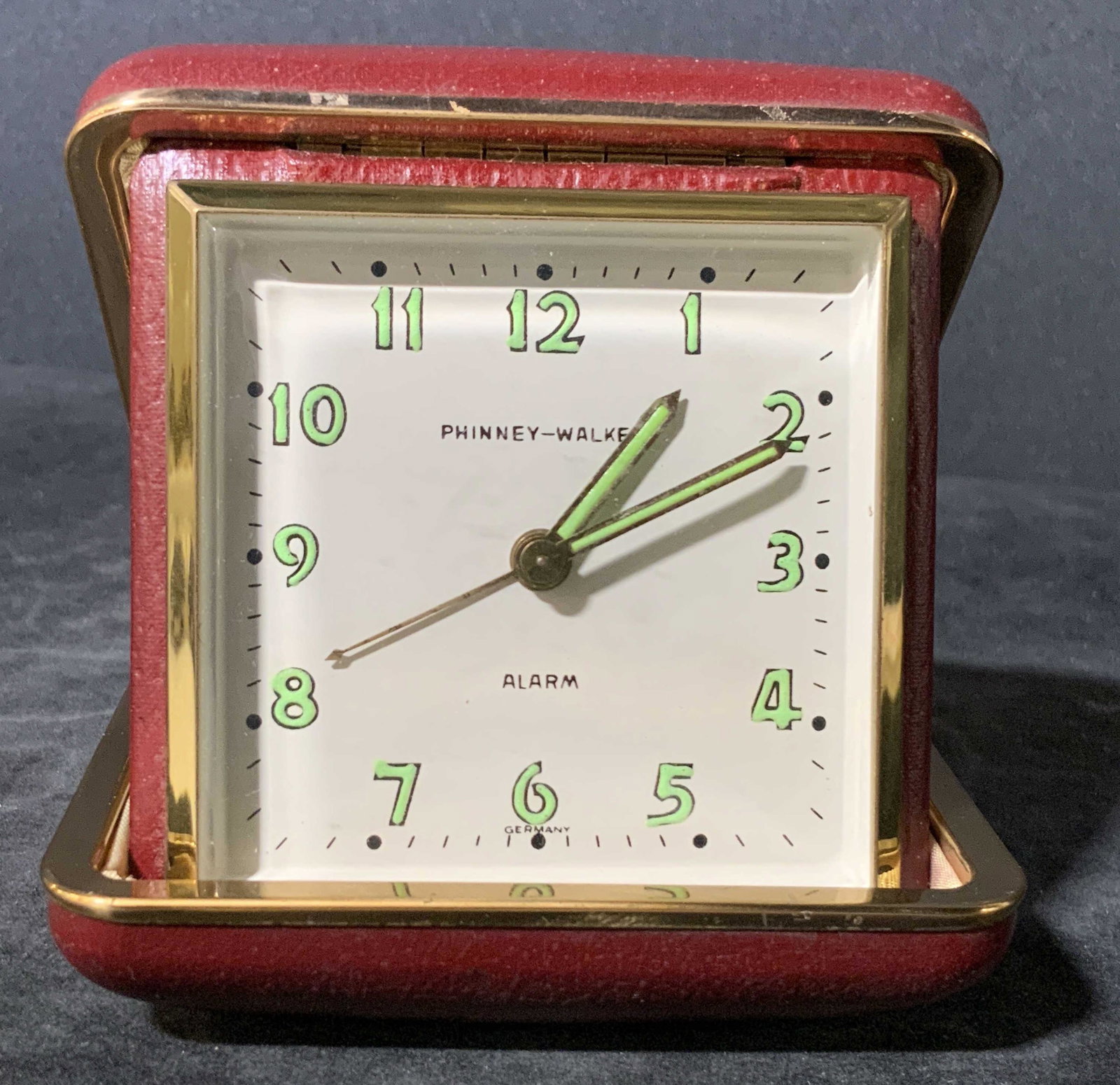 PHINNEY WALKER Foldable Alarm Clock In Case: A vintage Phinney Walker foldable clock. The piece exterior is a hard case, red hued with gold toned metal accents. Interior reveals clock. The clock is square in shape. Number clock face reads: PHINN