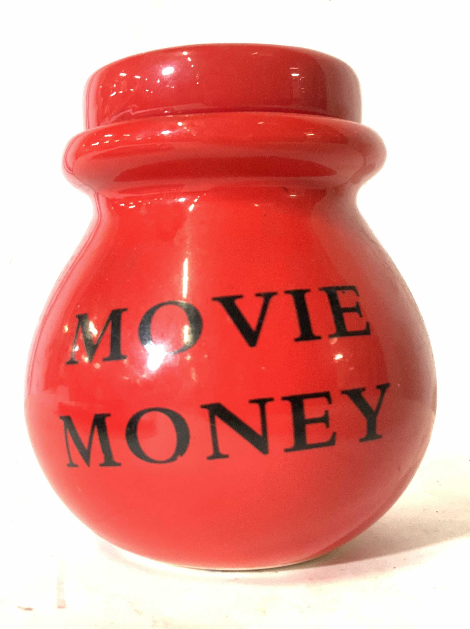 Vintage MOVIE MONEY Red Porcelain Coin Bank: A vintage red porcelain lidded coin bank. The frontside reads MOVIE MONEY. There is a slot at the top of the lid for inserting coins. Approx. 4.5H x 3W in. Property of Multi Generational Brooklyn Coll