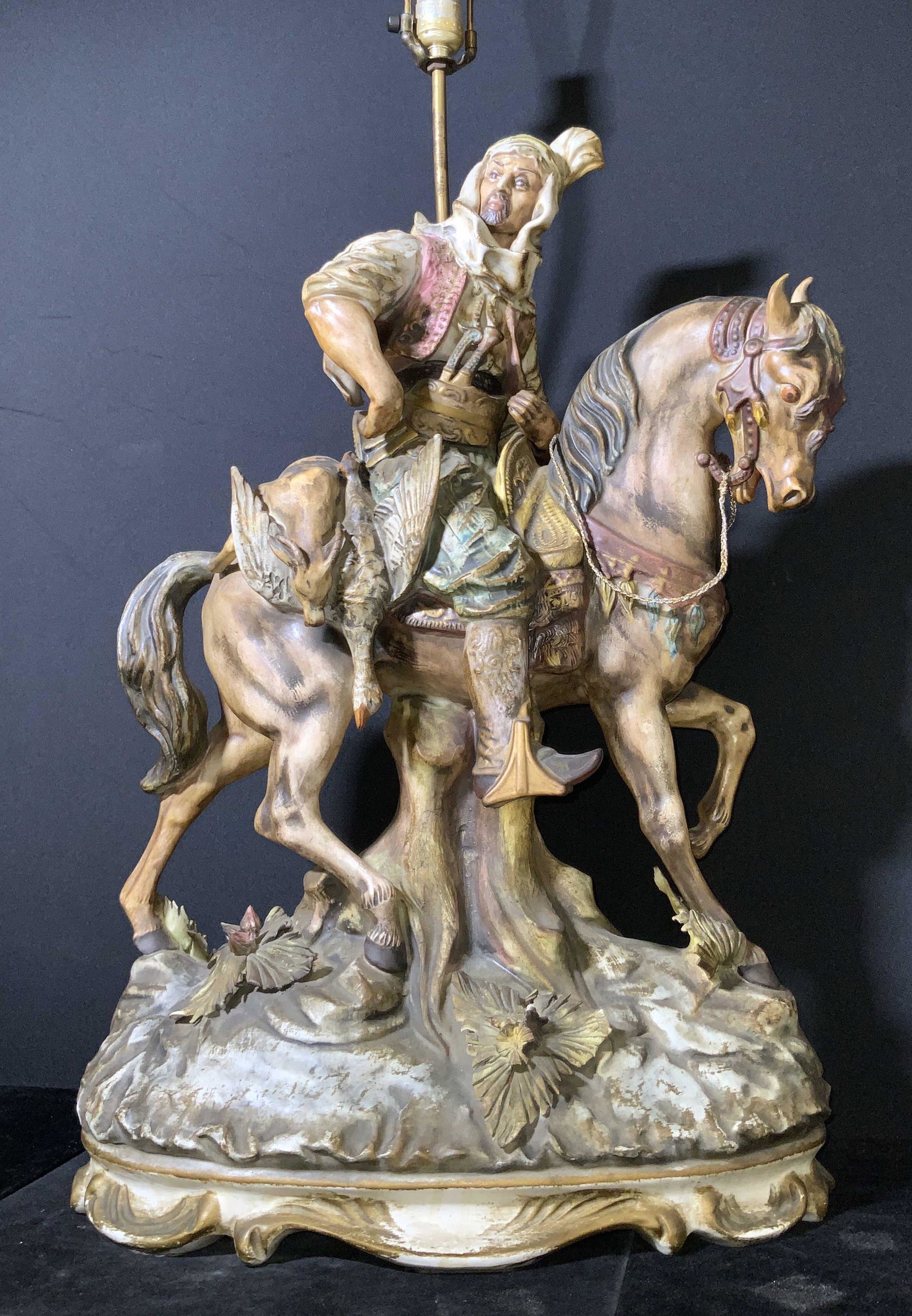Italian Porcelain & Metal Figural Tabletop Lamp: The piece depicts a man, a hunter, riding atop the back of a horse. A felled goose & deer are slung over the horse’s back. He wears a headdress and ornate clothing. Swords are tucked into his belt. 