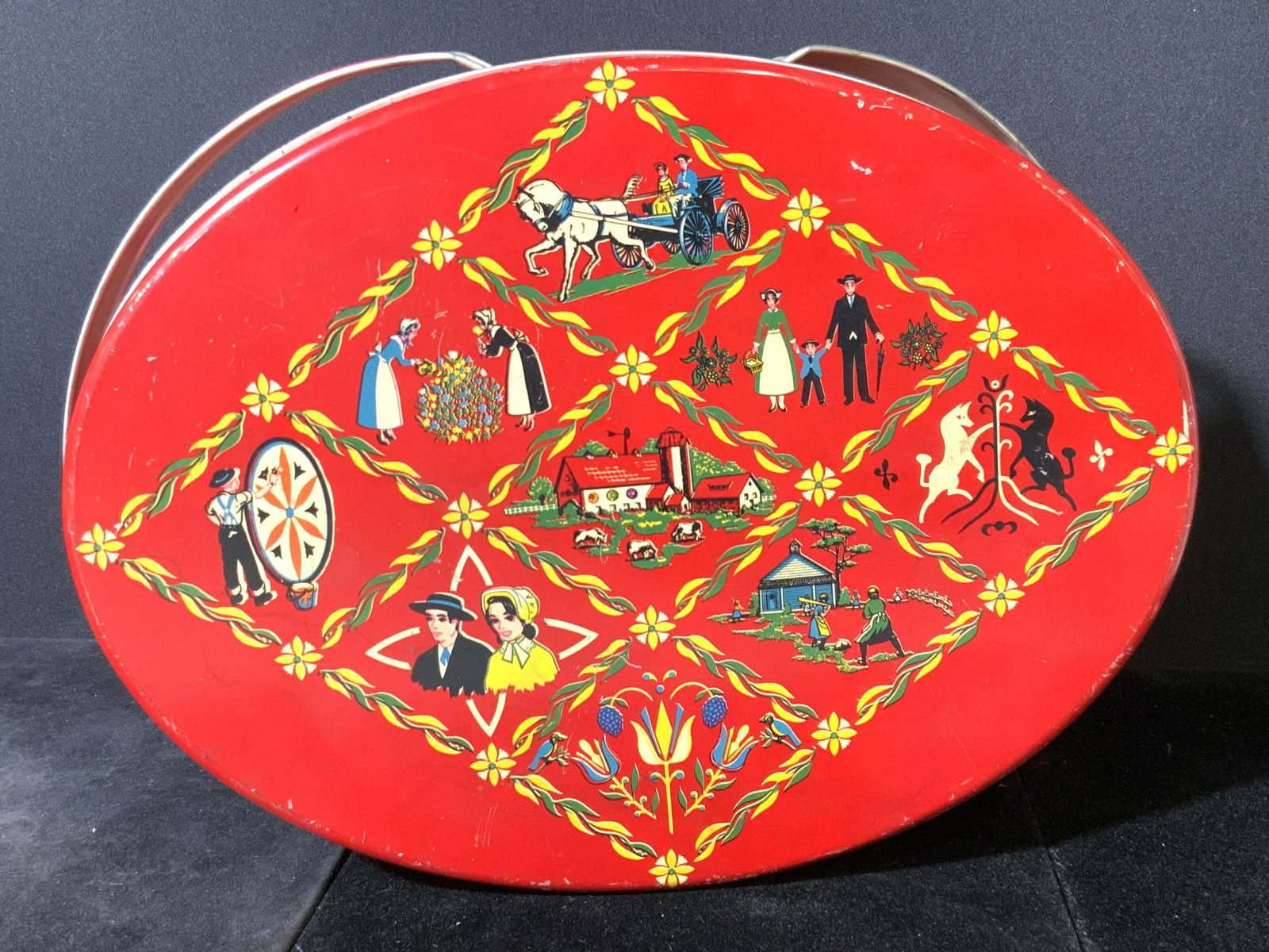 Vintage Collectible Red Folk Art Style Tin, Lid: A vintage red metal lidded basket tin. The exterior of the piece is a vibrant, retro red. The lid features folk art style depictions of people, horses, florals, homes, and more. There are two handles