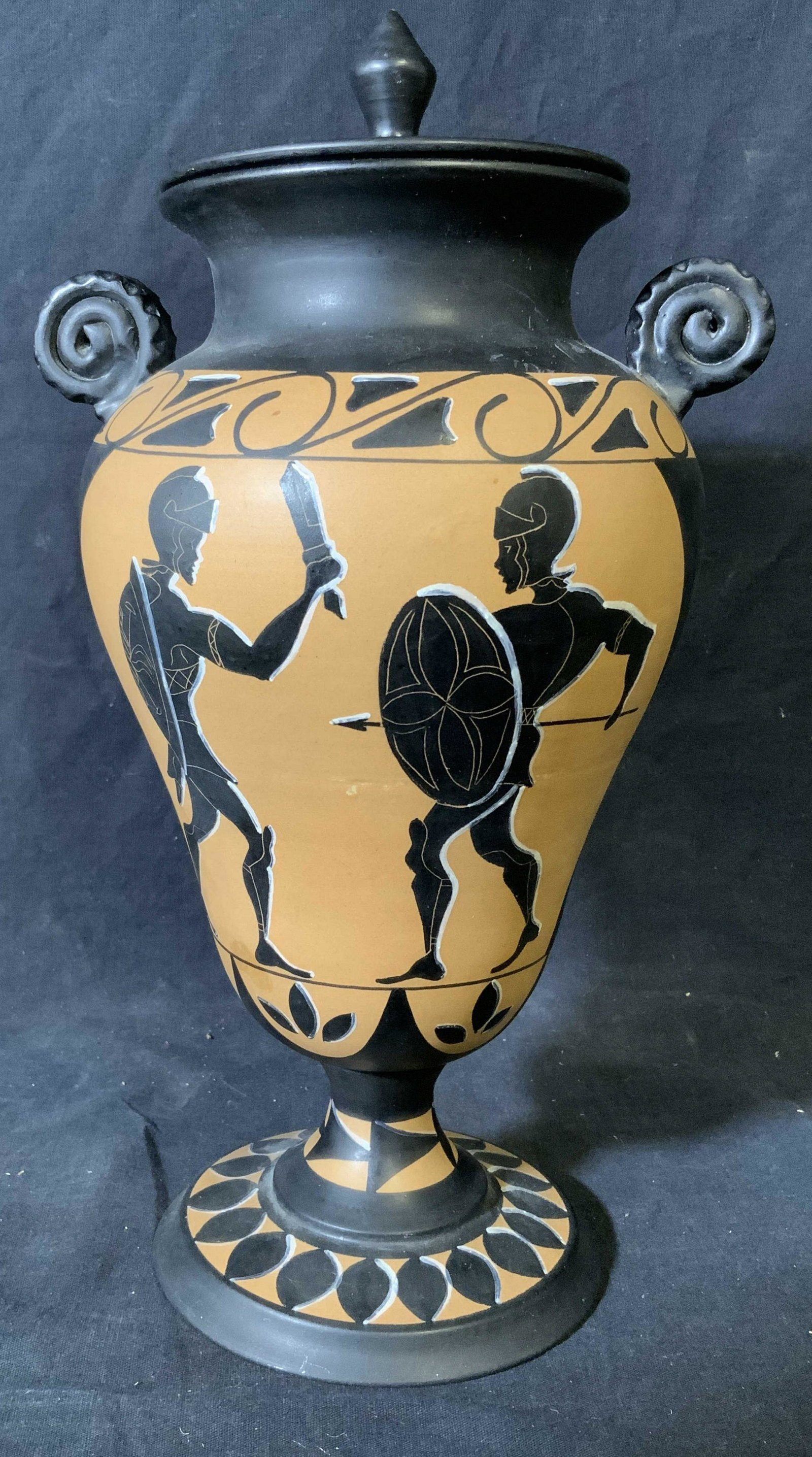 Hand Painted Grecian Style Ceramic Lidded Vase: Hand painted Grecian style ceramic lidded vase, label to underside reads ‘FH 58’, scene painted to surface depicts two soldiers fighting, piece painted in black white and brown, measures 13.5 inch