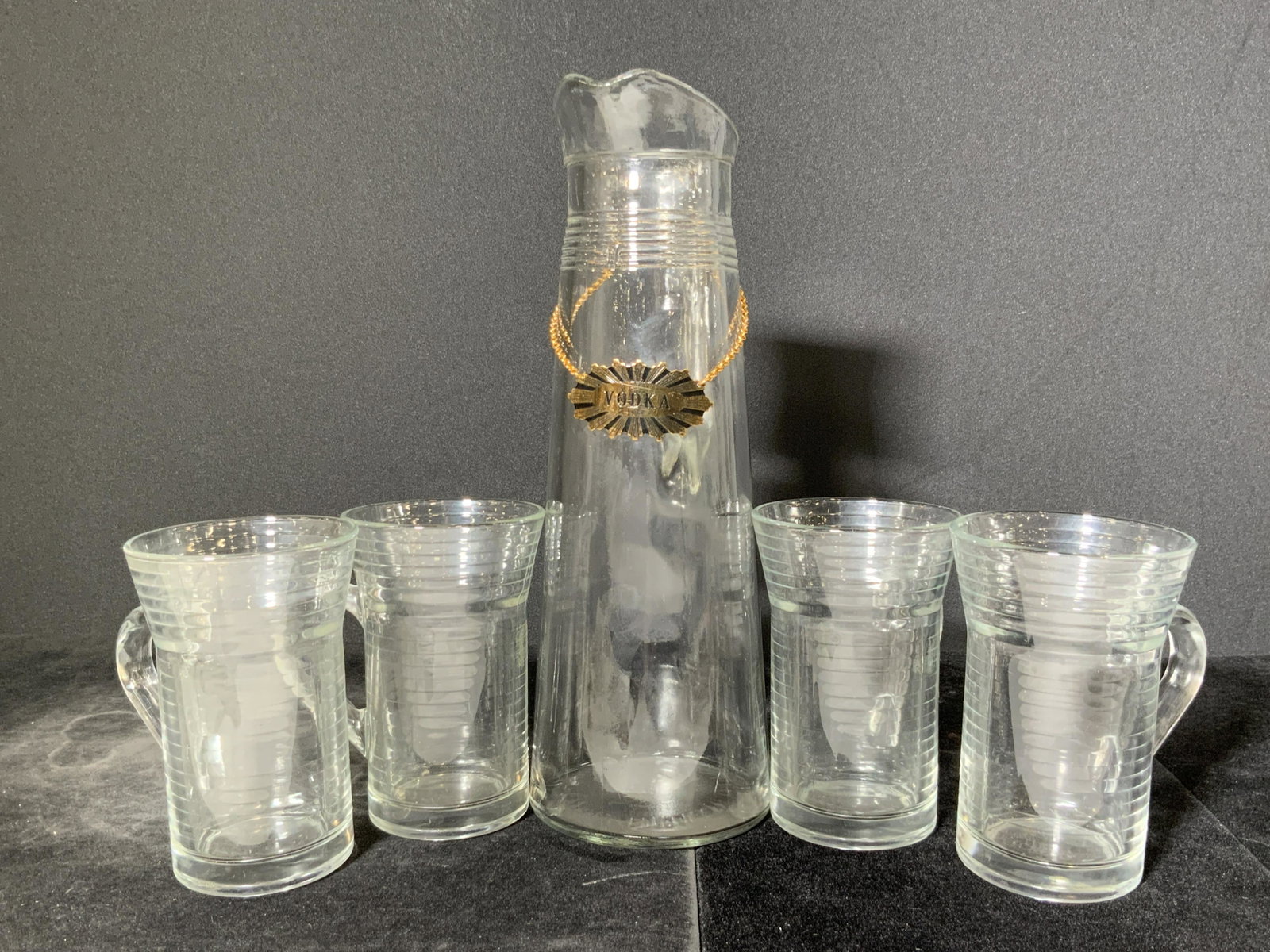 Set 5 Vintage Glass Cups & Pitcher, Barware: A set of four cups and one pitcher. Pieces are crafted from molded glass. Each piece is banded. Each piece has a handle. The pitcher has a gold tone chain wrapped around. The plaque at the center of t