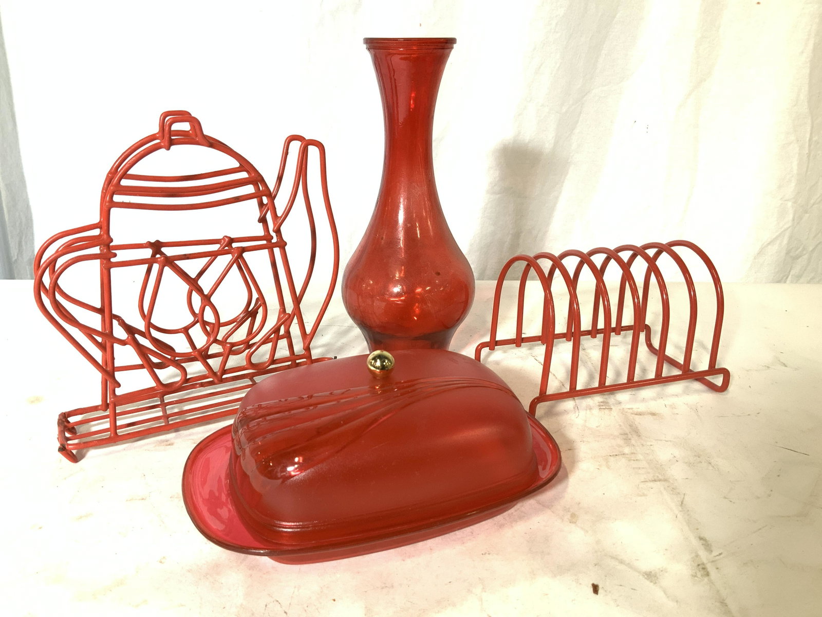 Lot 4 Red Tabletop Kitchenware: Coated red wire napkin holder in teapot form, toast separator, ruby red vase, plastic butter dish. Napkin holder measures 7 in. Tall. 6.75 in. L, has some wear to red coating. Property of Multi Genera