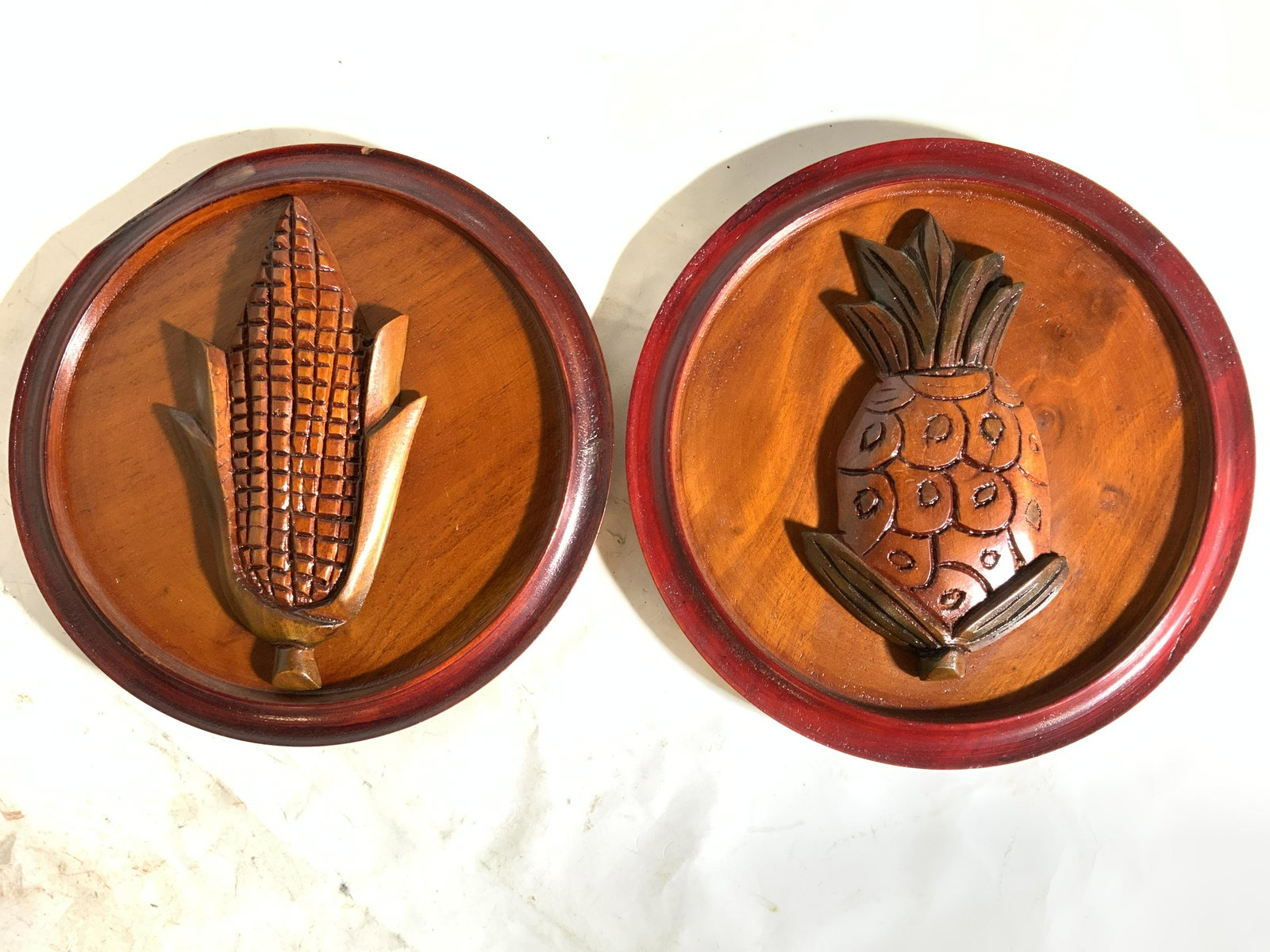 Lot 2 Grain & Fruit Wall Carvings: Each piece is possibly hand carved. One piece has carved image of corn, one has carved image of pineapple. Each have circular shape. Each measures approx 8.5 inches wide. Wall carvings, wall art, wood