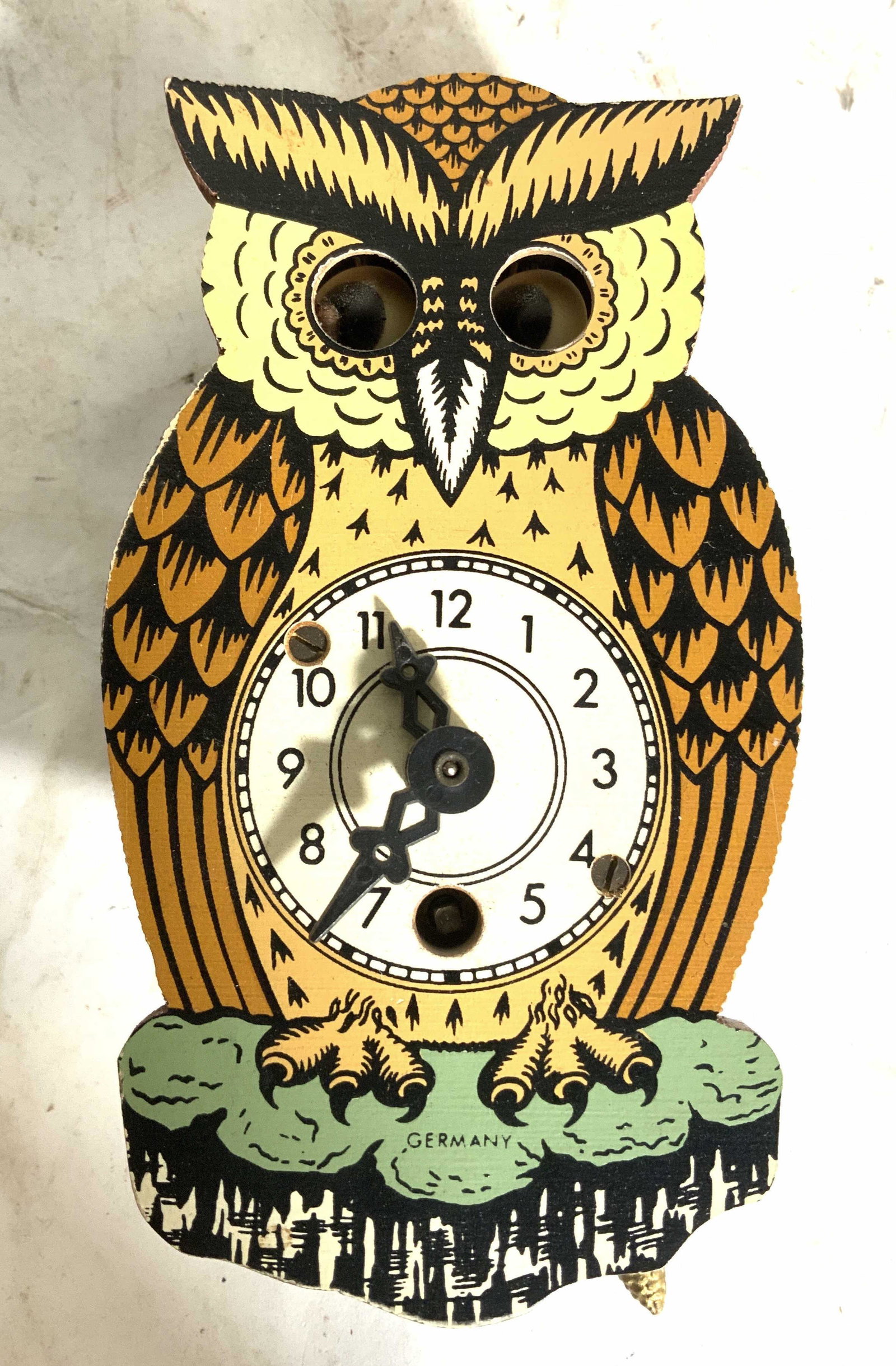 Petite Owl Shaped German Cuckoo Clock: Clock is petite in size. Front side reads GERMANY. Clock has 2 gold toned hanging Pinecone structures. Piece has black hand & numberswith cream toned clock face. Piece measures approx 5 inches tall 2.