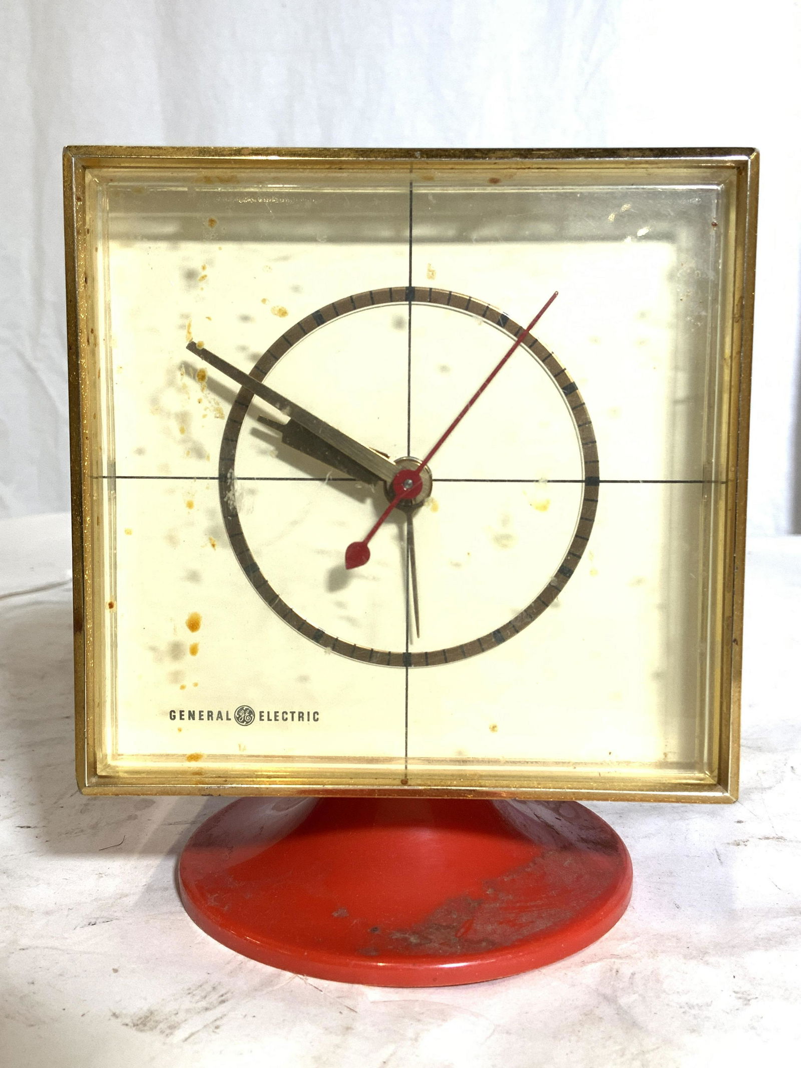 Vintage Retro GENERAL ELECTRIC Alarm Clock: Clock face reads GENERAL ELECTRIC. Clock is vintage with retro stylings. Piece is orange with gold toned framing around clock. Clock face is cream toned. Clock sits on pedestal stand. Piece when put t