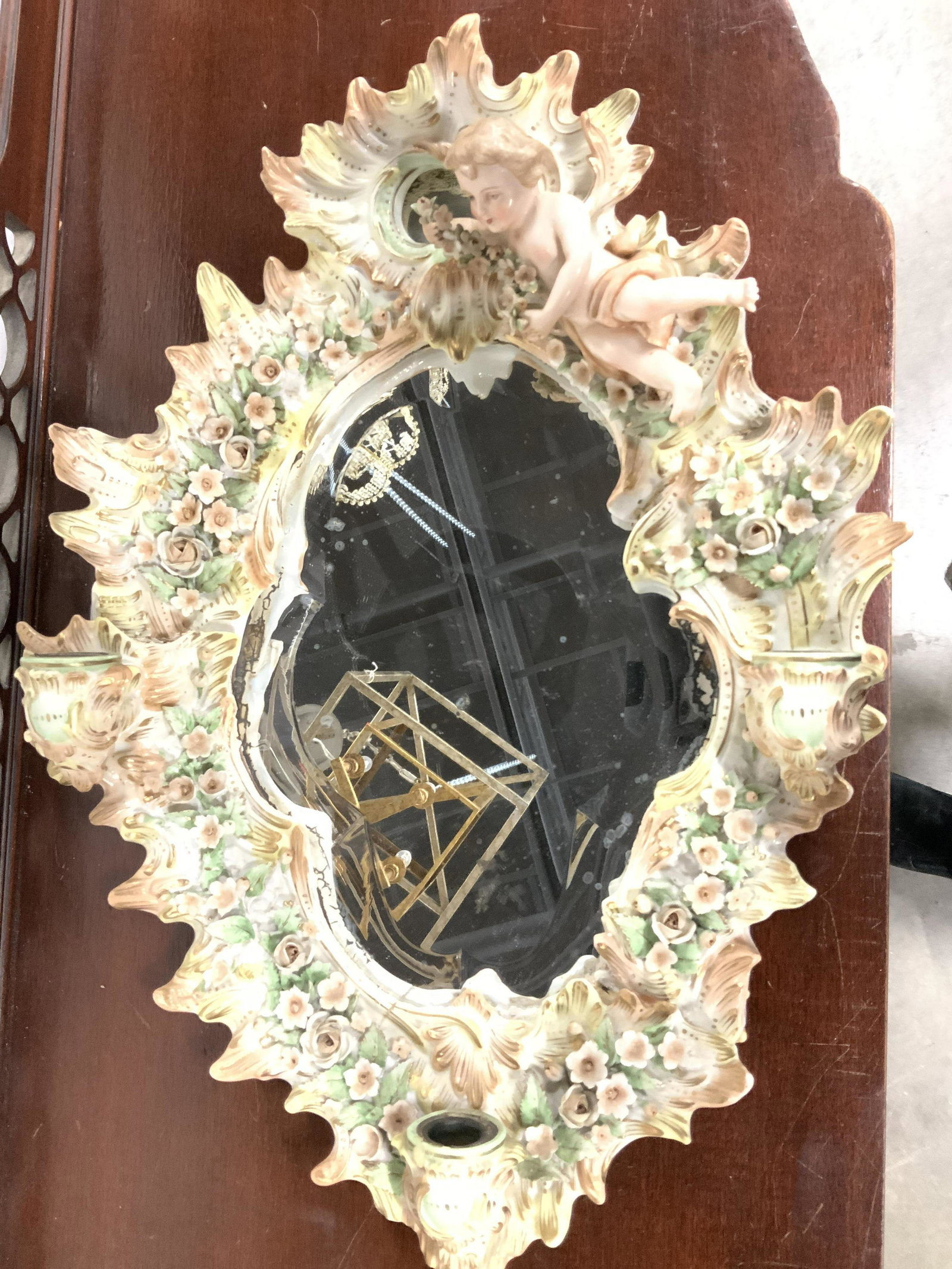 Antique Porcelain Cherub Venetian Style Mirror: Handmade gilt and hand painted pastels on white porcelain with flower and rose porcelain detail in relief. Large cherub putti holding floral vine, beveled mirror held by wood on backside. Hallmark in 