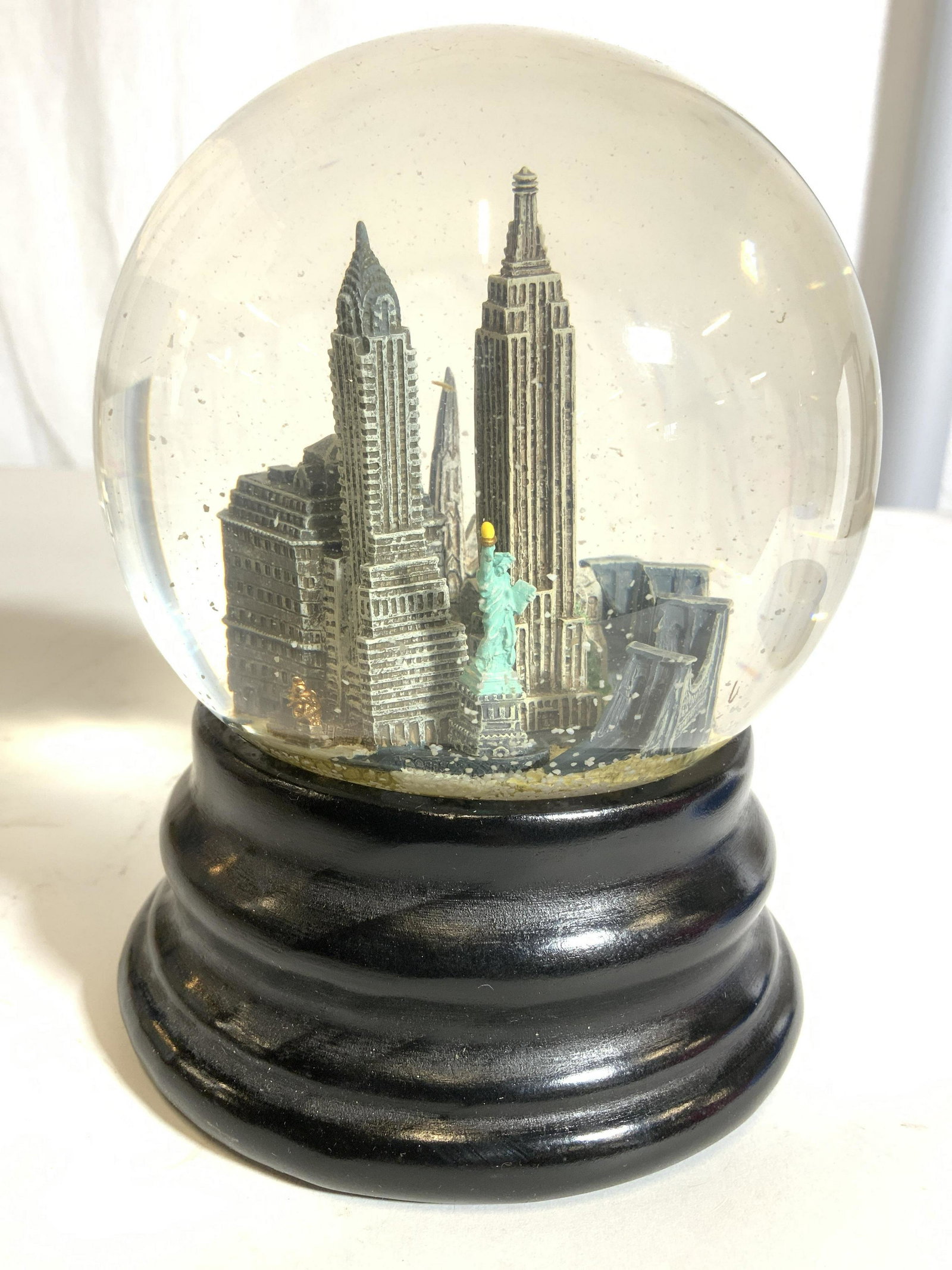 SAKS FIFTH AVENUE New York City Skyline Snowglobe: Underside of snow globe has tag which reads SAKS FIFTH AVENUE NEW YORK. Structure on interior resembles The New York City Skyline. Base is black toned and appears to be wooden. Piece measures approx 6