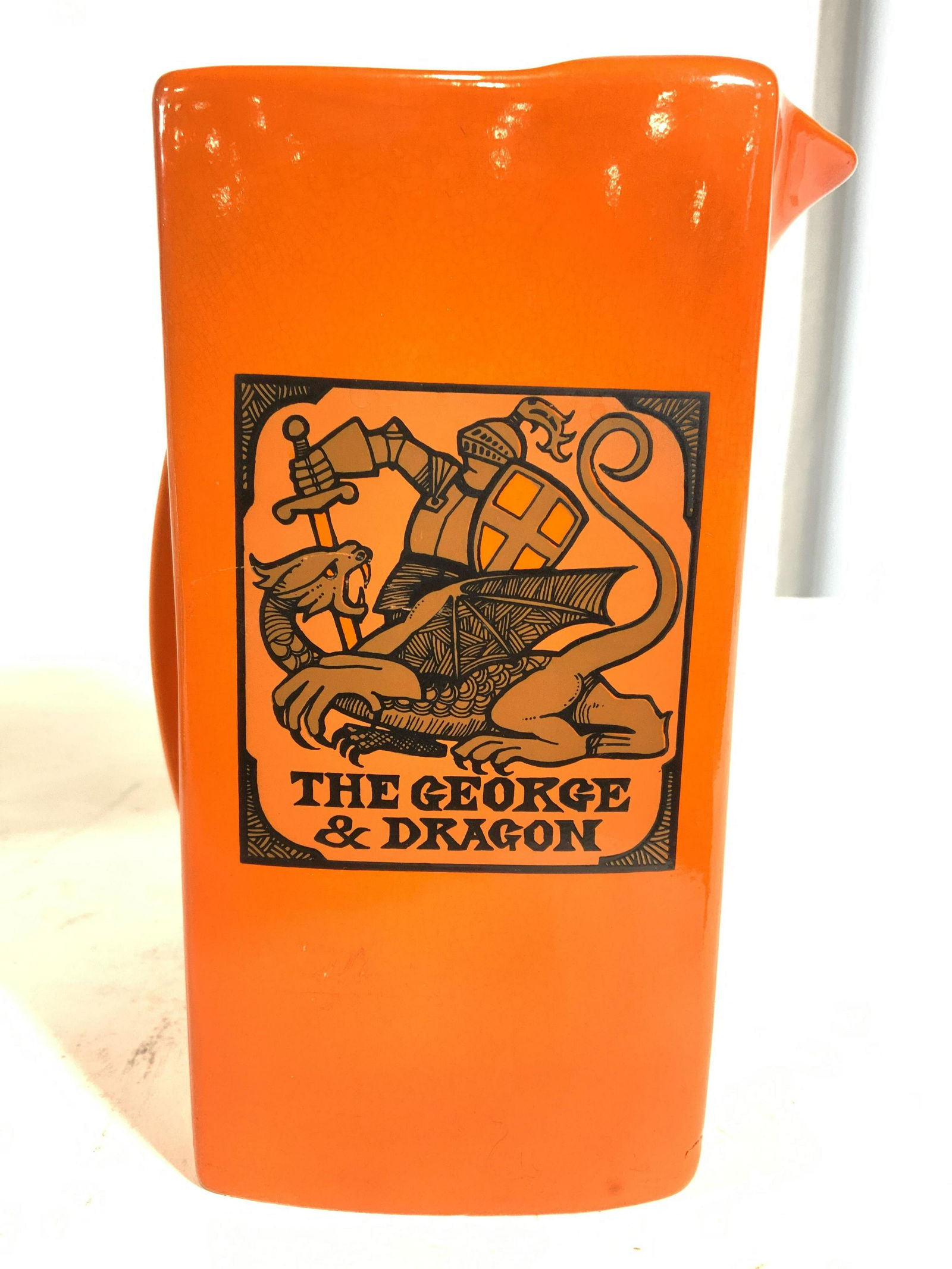 CARLTON WARE THE GEORGE & DRAGON Pitcher: Pitcher is porcelain. Piece has depict of and reads THE GEORGE & DRAGON on each side. Piece is orange & black toned. Piece measures approx 7.25 inches tall 5 inches deep 4 inches wide. Pitcher, porcel