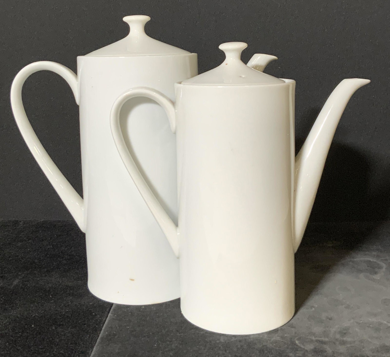 Pair Minimalist White Porcelain Lidded Pitchers: Two white porcelain pitchers with lids. These pieces have a minimalist design. One piece is larger than the other. Largest piece measures approx. 9H x 9W in. Some mild chips. Porcelain pitchers, poure
