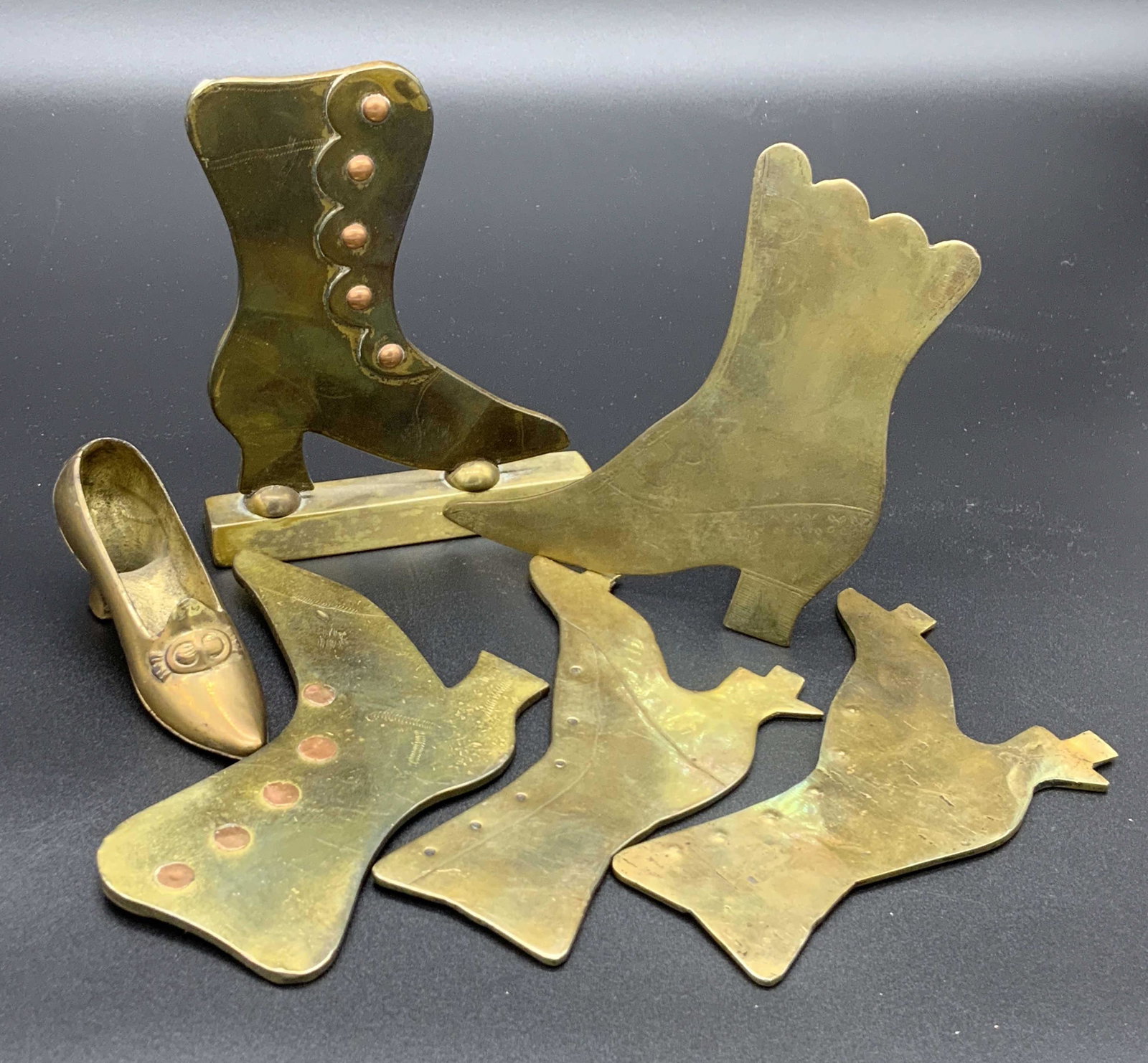 Lot 6 Brass Shoe Form Tabletop Accessories: Six pieces of polished brass accessories shaped like shoes. Some are standing, some lay flat, some pieces feature etched or embossed designs. Some wear to metals. Largest Approx. 4.5H x 3.5W in. Brass